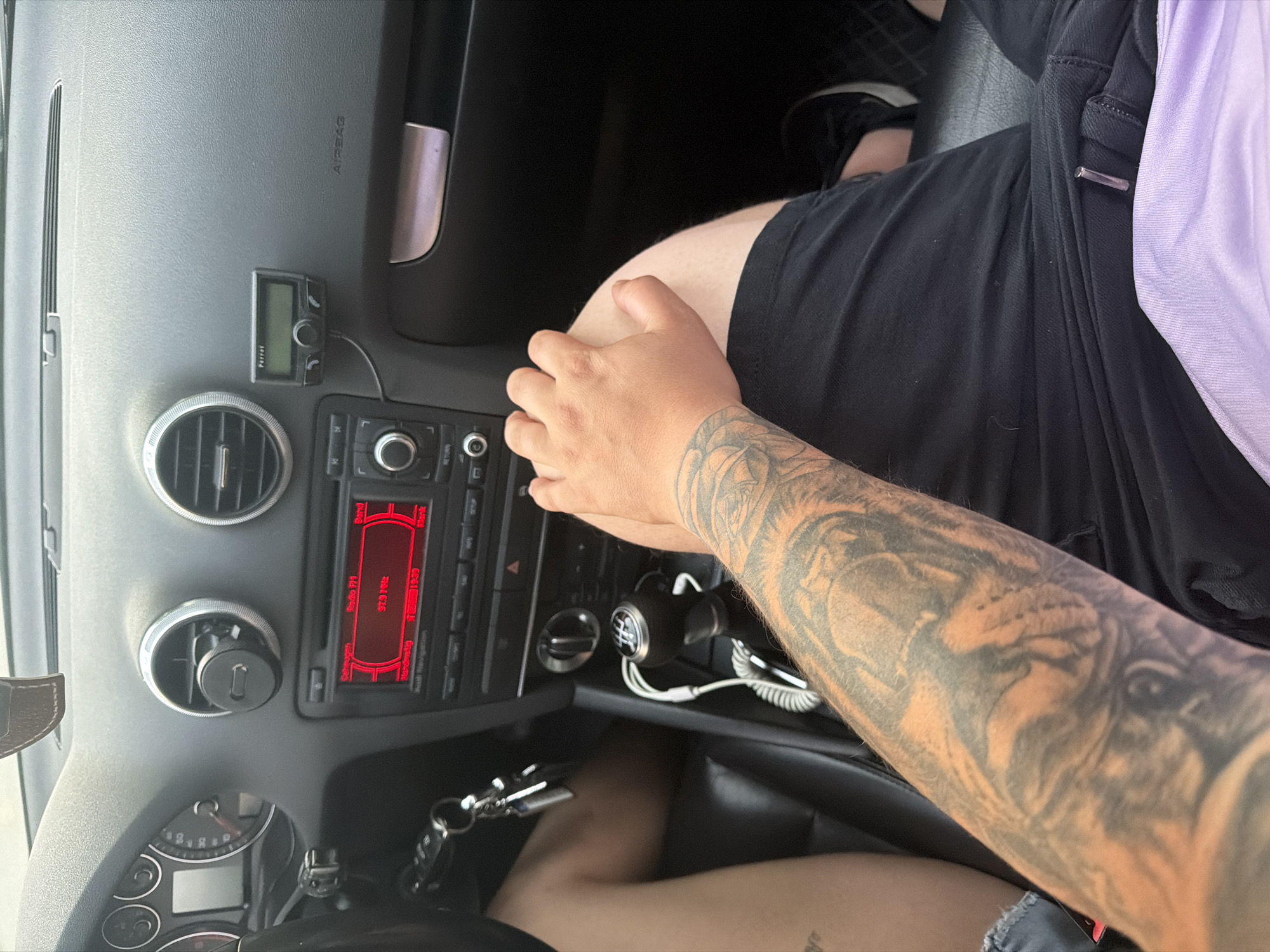 The image shows the interior of a car, likely an Audi A3 (8P generation), from a top-down perspective, with natural lighting suggesting daytime. A person is seated, visible from the torso down, wearing black shorts or pants and a light purple top. Their right forearm, heavily tattooed with an intricate design of a lion or large feline head, rests on their leg.
The dashboard features a radio unit with a red illuminated display showing "Radio FM" and "97.9 MHz," along with German text like "Senderwahl" (station selection), "Stau-Info" (traffic information), and "Navigationsinfo" (navigation information). Two circular air vents are visible on the dashboard, and a small black electronic device labeled "Parrot" is mounted above the left vent. The word "AIRBAG" is clearly visible on the passenger side dashboard. Below the center console, a manual gear stick is present, and a set of car keys hangs near the floor. The scene takes place inside a vehicle in Shumen, Bulgaria. The person appears to be a passenger or simply resting, with no specific activity in progress.
