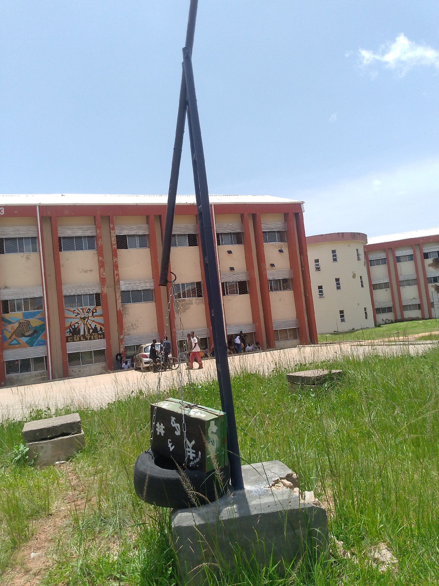 The image depicts an outdoor scene at Barakin Angundi, Nigeria. In the foreground, a metal pole with chains is anchored to a concrete base. Below the pole, a tire is suspended from chains, and a green box is hanging from the tire. The box has symbols of money, including the dollar, yen, and yuan signs, painted on it, along with the number three. Further back, a building with red trim and cream-colored walls is visible. The building has multiple floors with rows of windows. There are murals on the side of the building. Several people are gathered near the building, and cars are parked in front of it. The ground is covered with grass and dirt. The sky is blue with a few clouds.