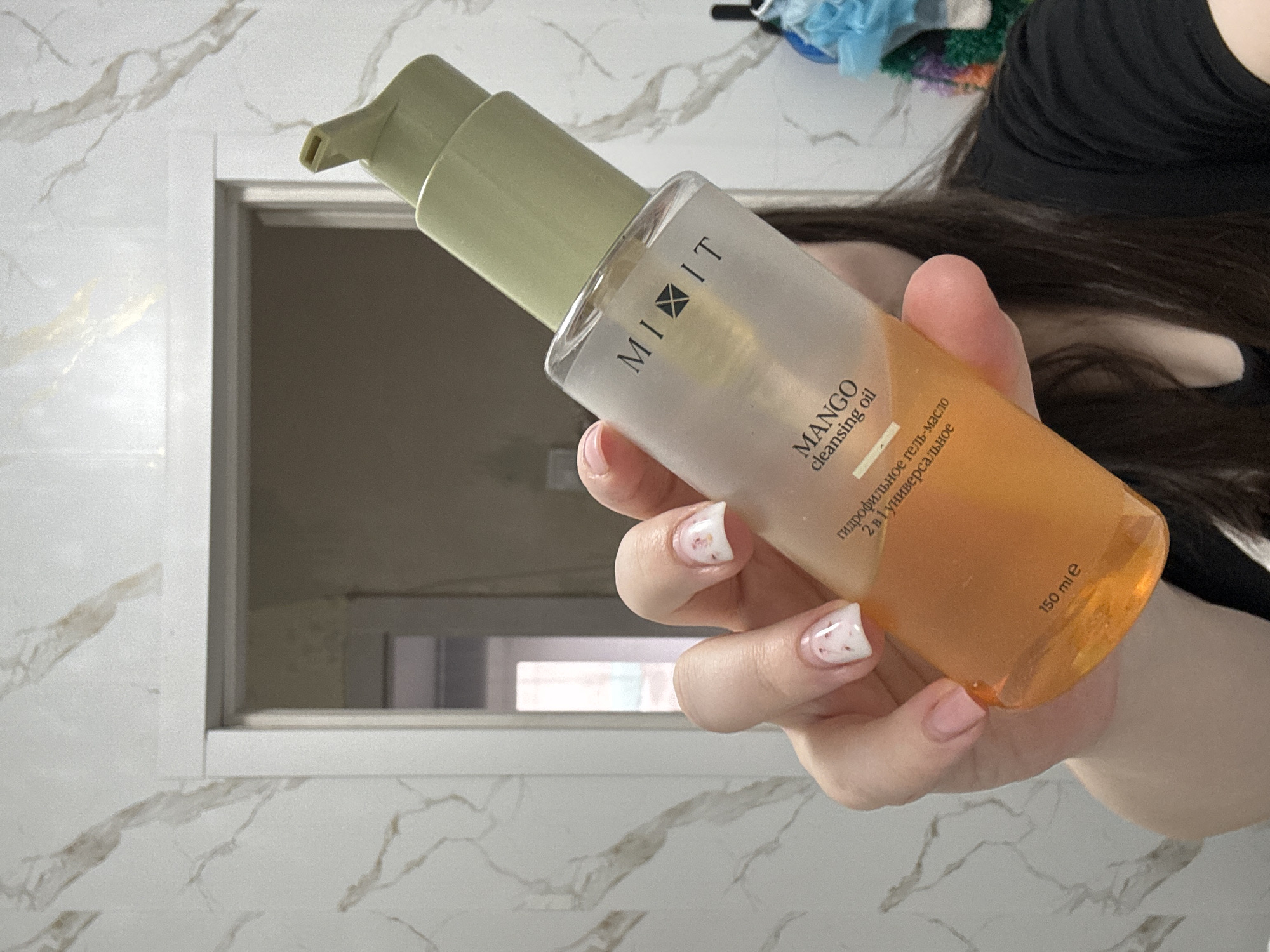 A hand holds a transparent bottle of "MIXIT MANGO cleansing oil" with a pale green pump. The bottle contains a gradient of liquid, pale on top and mango-colored at the bottom. The bottle's label contains text in both English and Russian. The hand has neatly manicured nails with a delicate floral design. The background features a white marble-patterned surface, a doorway or niche, and a glimpse of a person wearing a black shirt with dark hair. The location appears to be a bathroom, inferred from the materials and setting. The city of Chaikovsky, Russia, cannot be confirmed or refuted as the location based solely on the image.