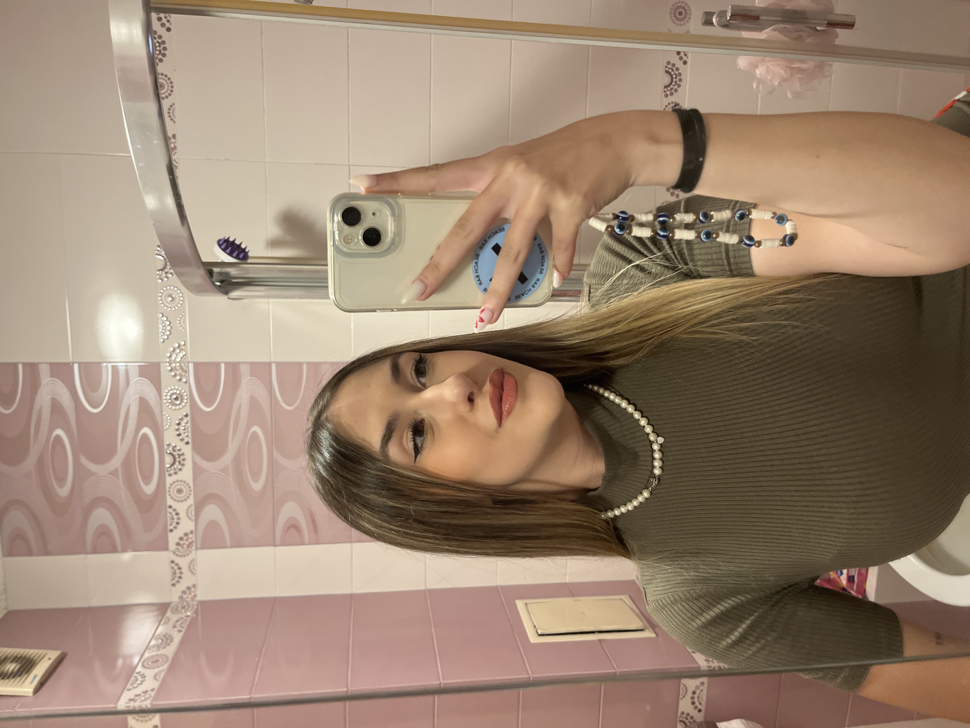 An indoor mirror selfie captures a young woman in a bathroom, likely located in Sofia, Bulgaria. She is shown from the chest up, with her long, straight brown hair flowing over her shoulders. She is wearing a form-fitting olive-green ribbed long-sleeve top, accessorized with a pearl necklace, a black bracelet on her right wrist, and a white beaded bracelet featuring several blue "evil eye" charms on her right forearm.
Her left hand holds a light-colored smartphone, possibly an iPhone, encased in a clear cover. On the back of the phone, a blue circular sticker is visible in reverse, displaying the text "BEACH DAYS." Her nails are long, manicured with white polish, and one finger has a small red heart design. The woman is wearing makeup, including visible eyeliner and lip gloss, and is looking towards her reflection.
The bathroom setting features pink and white tiled walls, with the white tiles adorned by a decorative border of circular patterns. Various bathroom items are reflected in the mirror: a metal shower caddy containing a purple, spiky object (possibly a scalp massager), a white toilet in the lower right, and a pink loofah hanging from a rail in the background. A small comb is also visible on a surface in the lower left. The lighting appears artificial, consistent with an indoor bathroom setting.