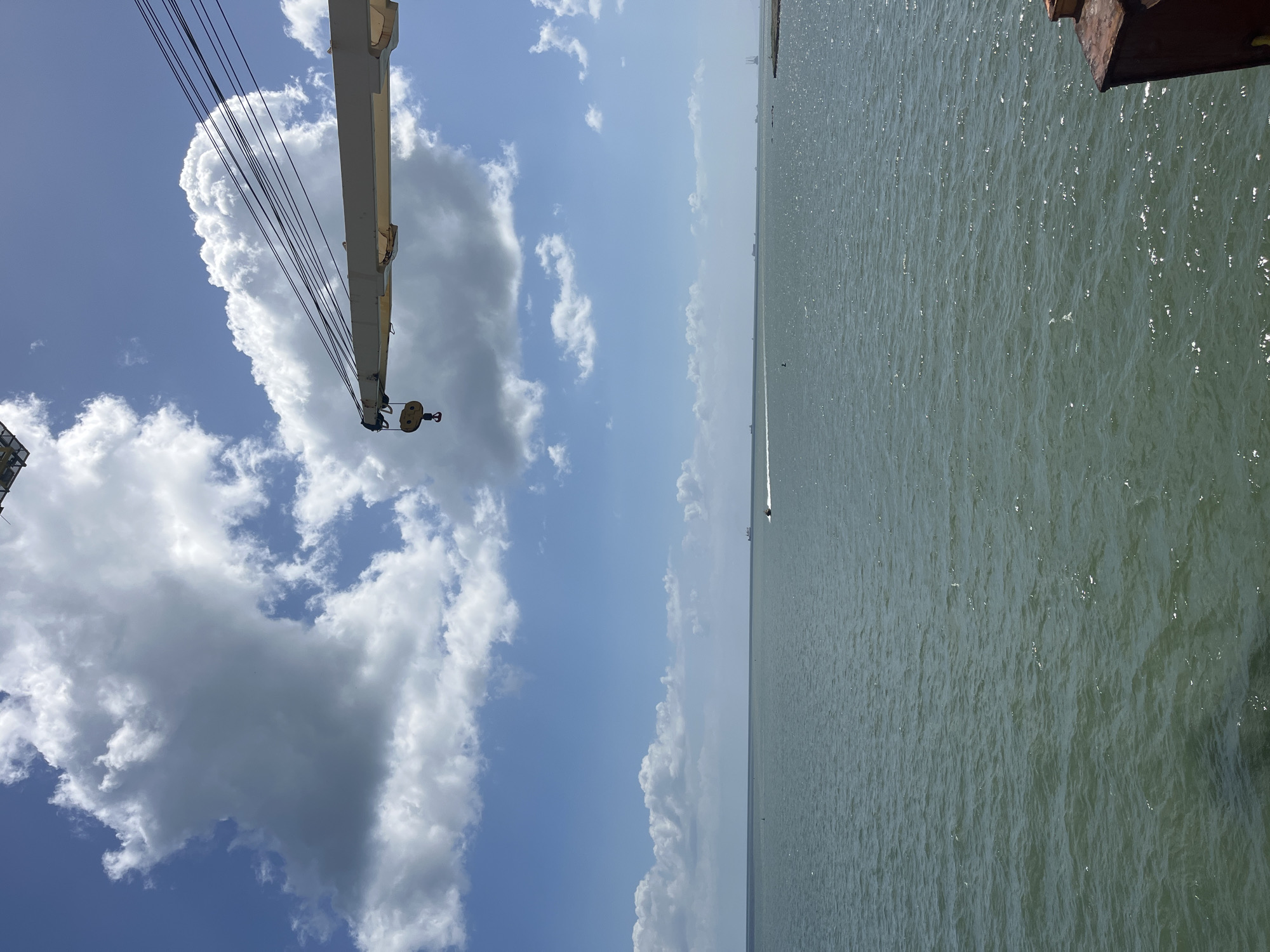 The image displays a bright, daytime scene primarily composed of water and sky, featuring industrial elements. A vast body of water, likely a sea or bay, occupies the lower half of the frame, extending to a distant, hazy horizon line. The water's surface is lightly rippled, with numerous sunlit glints indicating calm to moderate conditions.
Above the water, a clear blue sky is filled with scattered, white cumulus clouds of varying sizes and shapes. In the upper left quadrant, the prominent, light-colored boom of a large crane extends diagonally downwards, cutting across the sky. Its intricate system of dark cables and a yellow hook block with a single pulley are clearly visible near the center of the frame.
In the far distance on the water, a small white boat can be discerned, providing a sense of scale and depth. A dark, angular structure, possibly a section of a dock, pier, or another vessel, is partially visible in the upper right corner.
The overall impression is one of a sunny, pleasant day. There are no people visible, and no specific event or activity is taking place beyond the presence of the boat. No text is present in the image. The setting suggests a coastal or port environment.