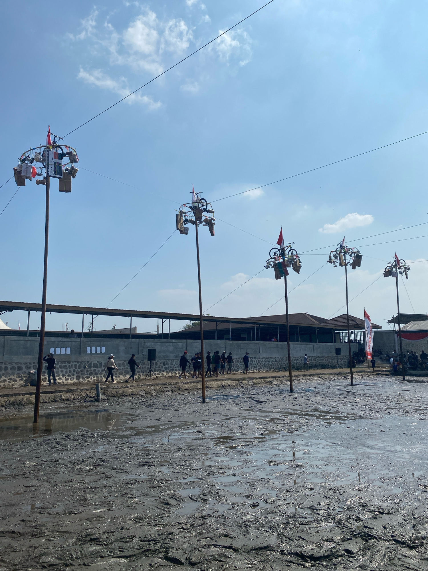 The image depicts a muddy field with several tall poles adorned with decorations and flags, suggesting a festive or ceremonial event. The poles are positioned in a line across the muddy terrain, with some carrying what appear to be miniature bicycles and various colorful items, possibly prizes or offerings. In the background, a low wall separates the field from a row of buildings, and a crowd of people are visible observing the scene. The sky is clear and blue with scattered clouds. The location is identified as the "Lapangan Gajahmada" in Kediri City, Indonesia. The presence of numerous poles with decorations and a gathering of people in a muddy area indicates a local celebration or competition, possibly related to Indonesian Independence Day given the red and white flags.