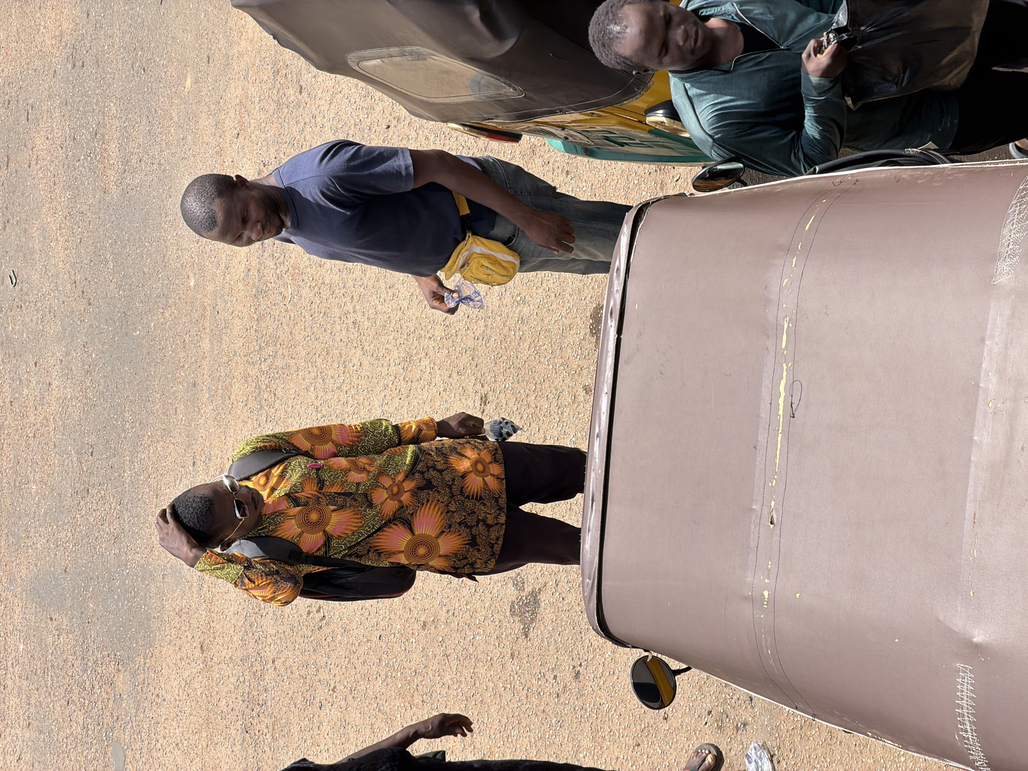 In Bukuru, Nigeria, two men stand on a dirt road next to a three-wheeled vehicle. The man on the left wears a brightly colored shirt with a floral pattern, sunglasses, and a backpack. He is touching his head. The man on the right wears a navy blue t-shirt and jeans, and has a yellow fanny pack around his waist. He is holding a clear plastic bag. Another three-wheeled vehicle is visible to the right, partially obscuring the view. A third man in a green jacket is standing next to the vehicles on the right side of the image. The road is made of gravel and dirt, and the lighting suggests it is daytime.