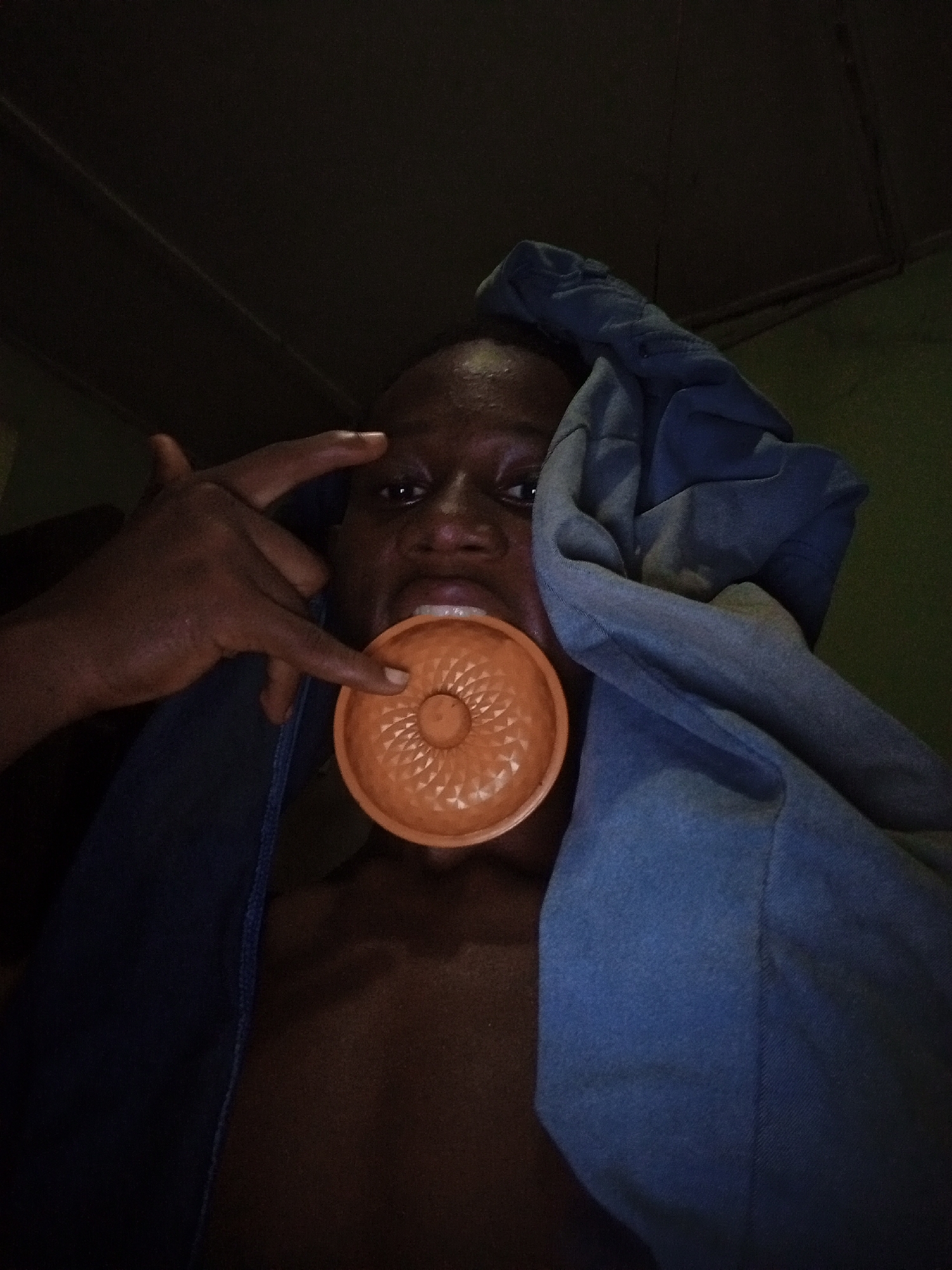 A low-angle shot depicts a dark-skinned person holding an orange, circular object with a raised, patterned center in their mouth. The person points to the object with a finger from their left hand. A blue fabric is draped around their upper body and head. The environment appears dimly lit and green. The location is stated as Ukwu-Abwa, Nigeria, but this is not visually verifiable from the image.