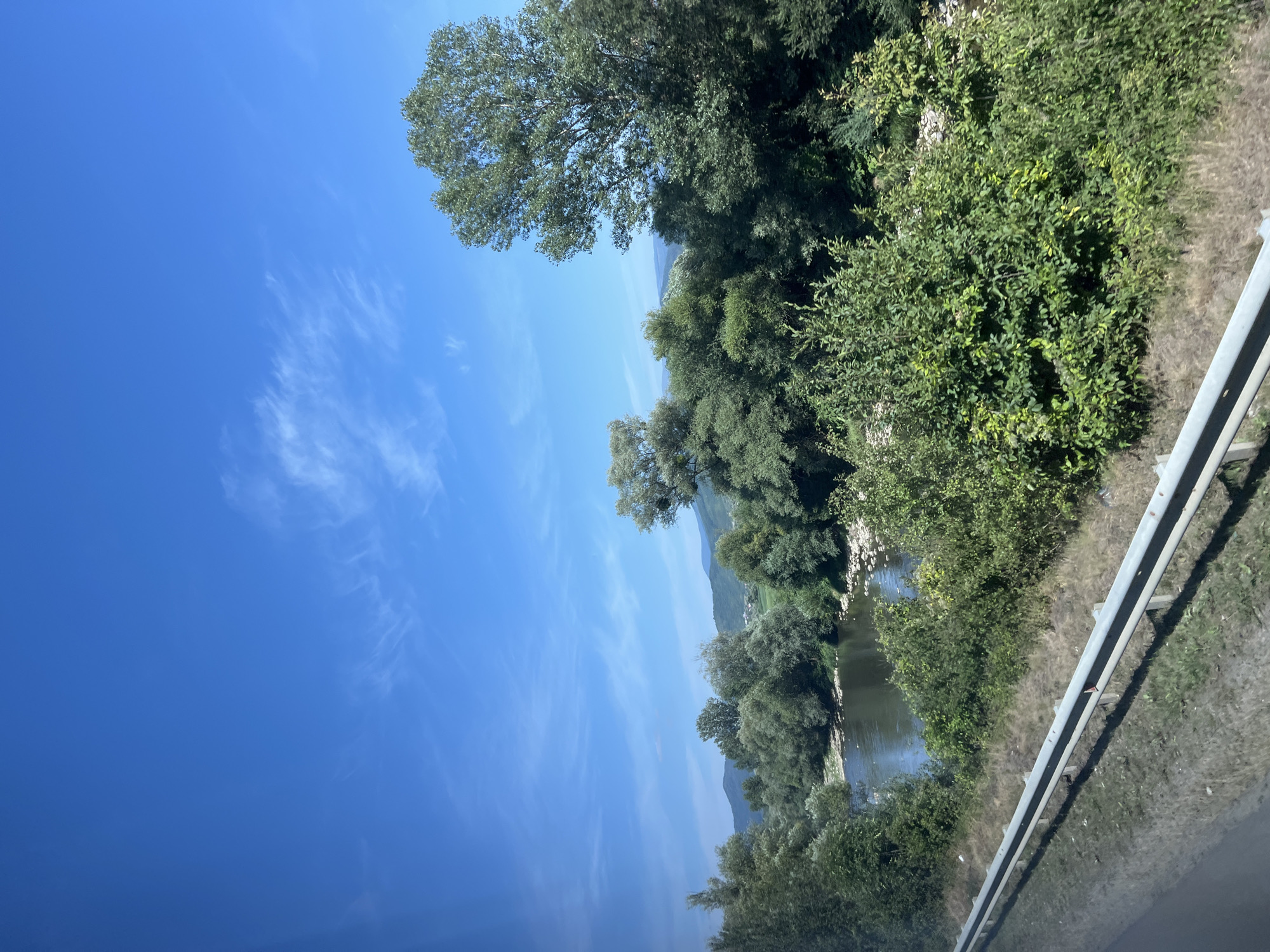 The image shows a scenic view of a river flowing through a landscape with lush green trees and vegetation. In the foreground, a metal guardrail runs along the side of what appears to be a road. The river is calm, with rocky banks visible on the far side. The background features rolling hills covered in dense foliage under a bright blue sky with scattered wispy clouds. The overall impression is of a natural, serene environment.