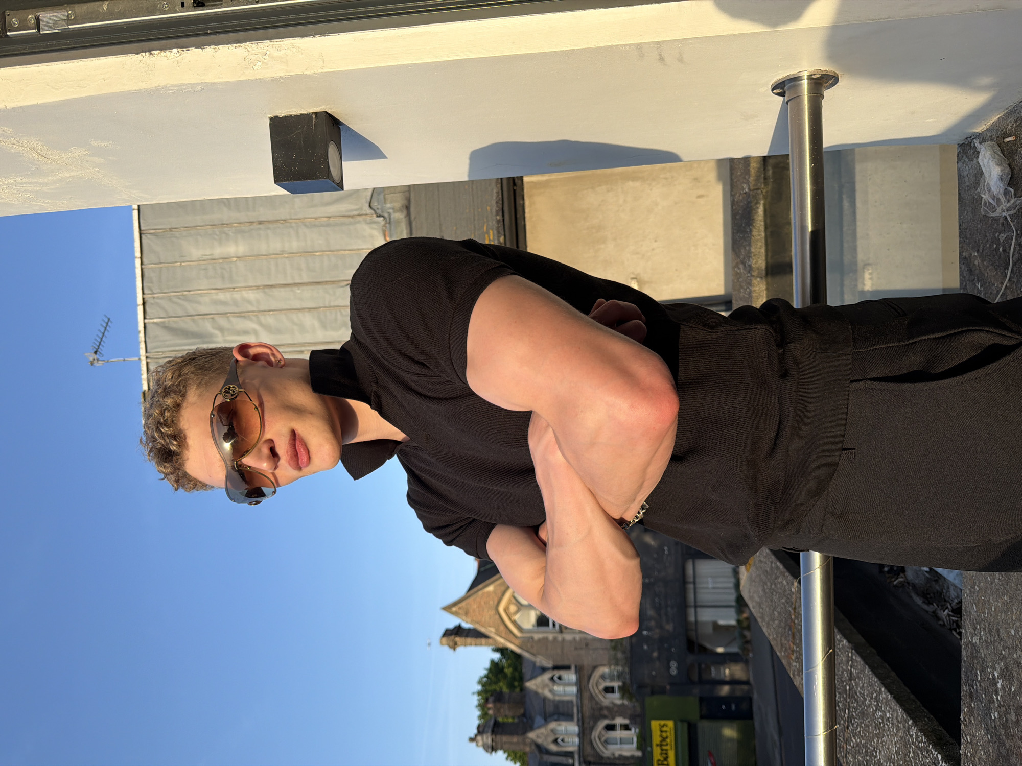 A man stands outdoors, likely on a balcony, with a building in the background identified by a sign reading "Barbers", suggesting a location in London, United Kingdom. He is wearing sunglasses and a dark-colored outfit. His arms are crossed. The scene is captured in bright daylight under a clear blue sky. A metal railing is visible beside the man, along with a speaker attached to the roof. An antenna can be seen far in the distance.