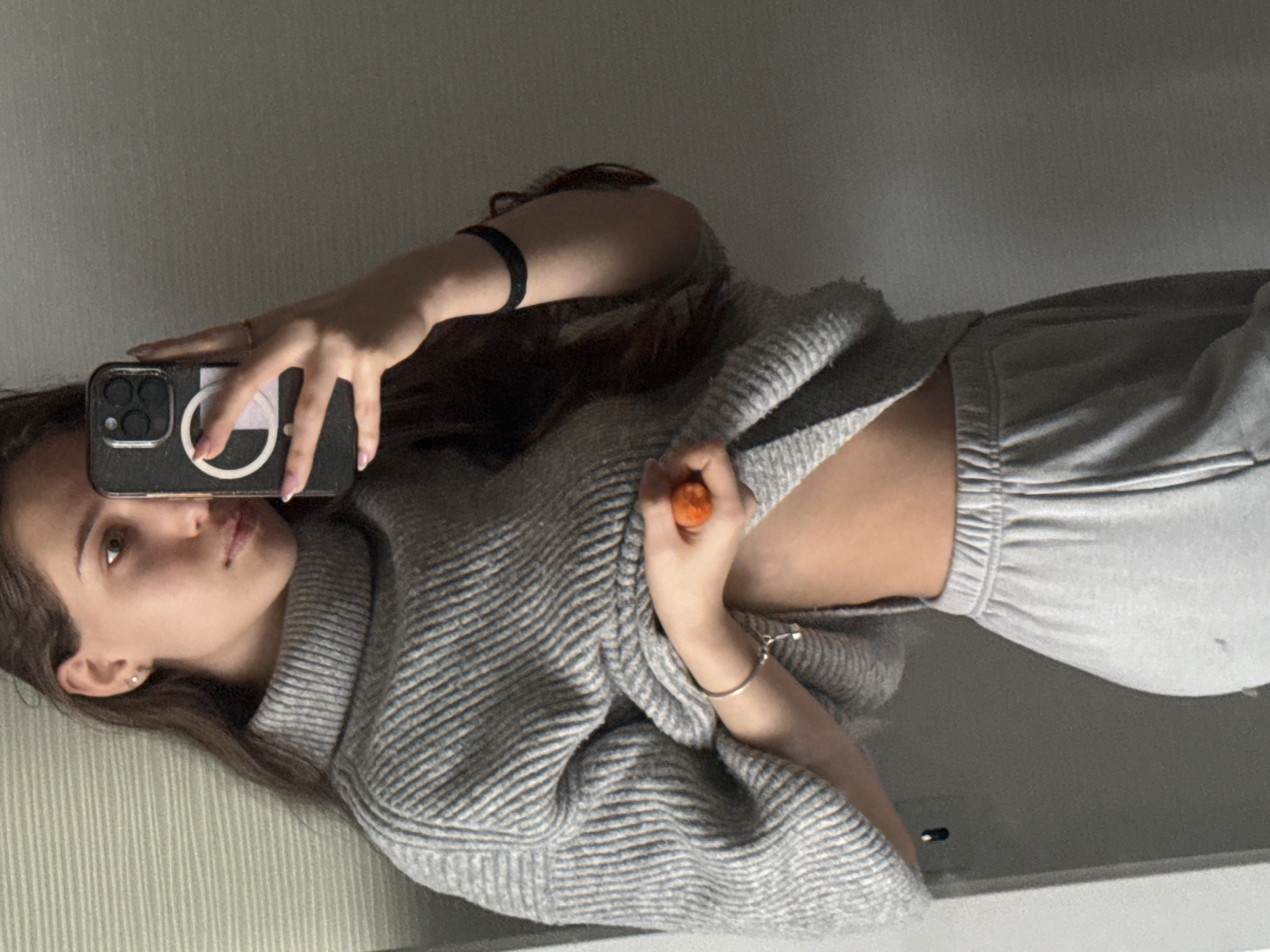 A woman is taking a selfie in what appears to be an indoor setting. She is holding her phone up to her face, partially obscuring it. The woman is wearing a gray, textured turtleneck sweater that is slightly pulled up on one side, revealing a portion of her abdomen. She is also wearing gray sweatpants. There are two bracelets on her left wrist, and an orange object is visible near her hand. The background is a plain, neutral-colored wall. There is no indication of the location being Sofia, Bulgaria.