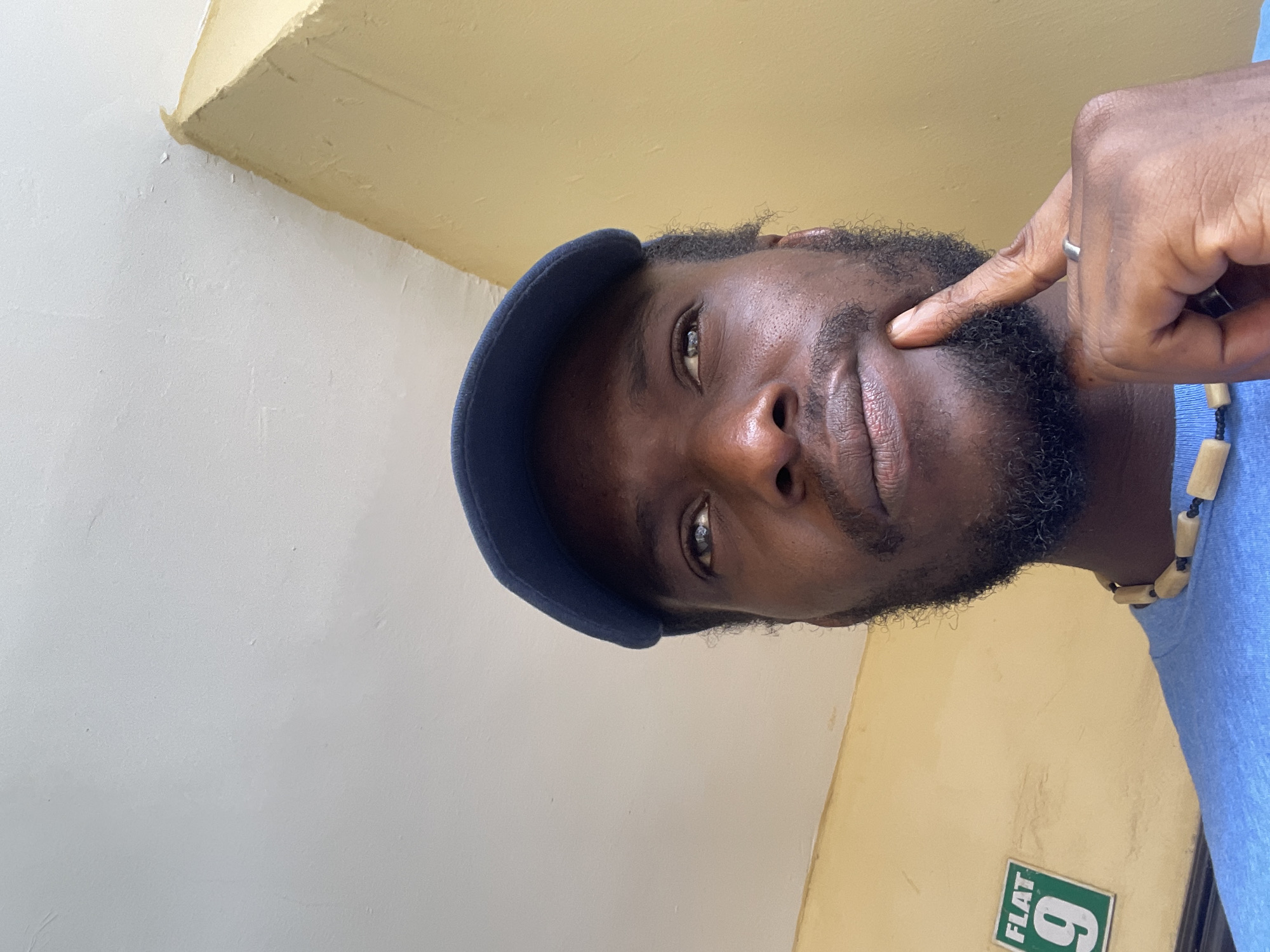 A close-up, low-angle, outdoor shot captures a Black man wearing a blue baseball cap and a blue t-shirt, adorned with a wooden bead necklace, as he rests his index finger on his chin in a pensive gesture. He has a neatly trimmed beard and his gaze is directed upwards, as if in thought. To his left, a green and white sign indicates "FLAT 9". The background consists of a yellow wall meeting a white ceiling, with a hint of a corner where the two surfaces join. The setting is likely a residential area in Abuja, Nigeria, given the presence of a flat number sign.