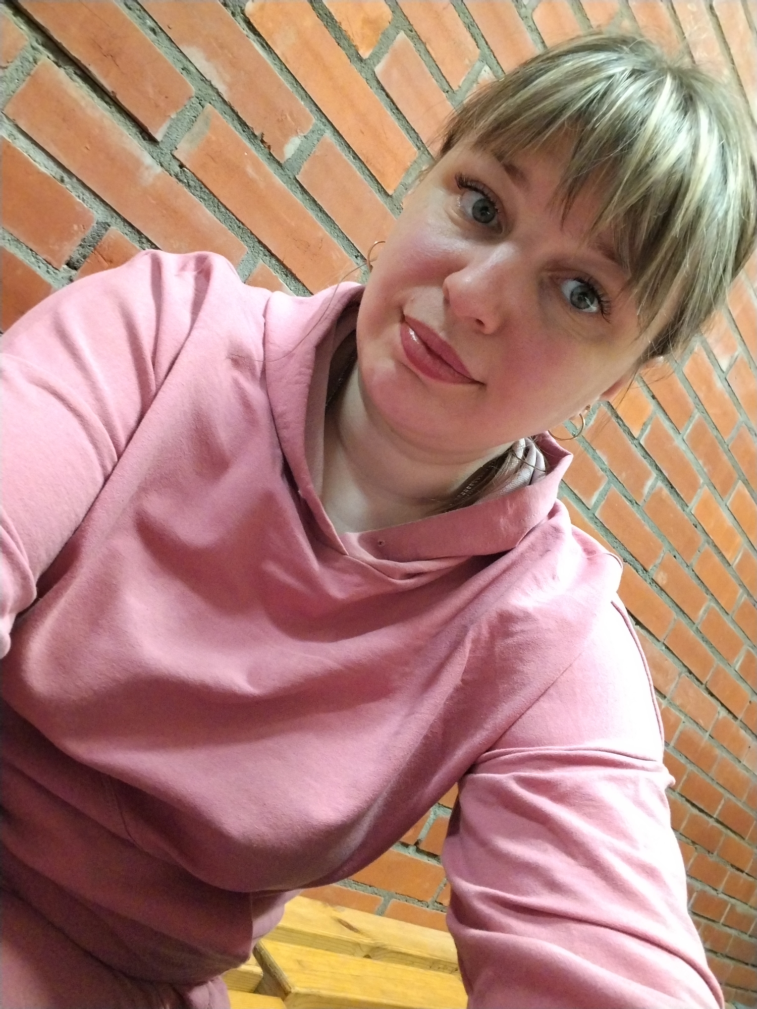 A medium close-up, slightly high-angle selfie features a woman with light-colored hair and bangs, looking directly at the camera with a gentle, inviting expression. She has wide, light-colored eyes (likely blue or grey), defined eyelashes, and her lips are slightly parted, covered in a glossy finish. Her hair is a mix of blonde highlights and darker roots, swept to frame her face.
She is wearing a pink long-sleeved hooded sweatshirt, which dominates the lower half of the frame with its soft fabric and subtle folds. A glimpse of a small gold hoop earring is visible on her right ear. The background consists of a rustic-looking brick wall, with reddish-orange bricks laid in a diagonal pattern and contrasted by grey mortar lines. Below her, a portion of a wooden surface, possibly a bench or furniture, is visible, suggesting an indoor or sheltered outdoor setting. There is no visible text in the image. The overall mood is casual and direct.