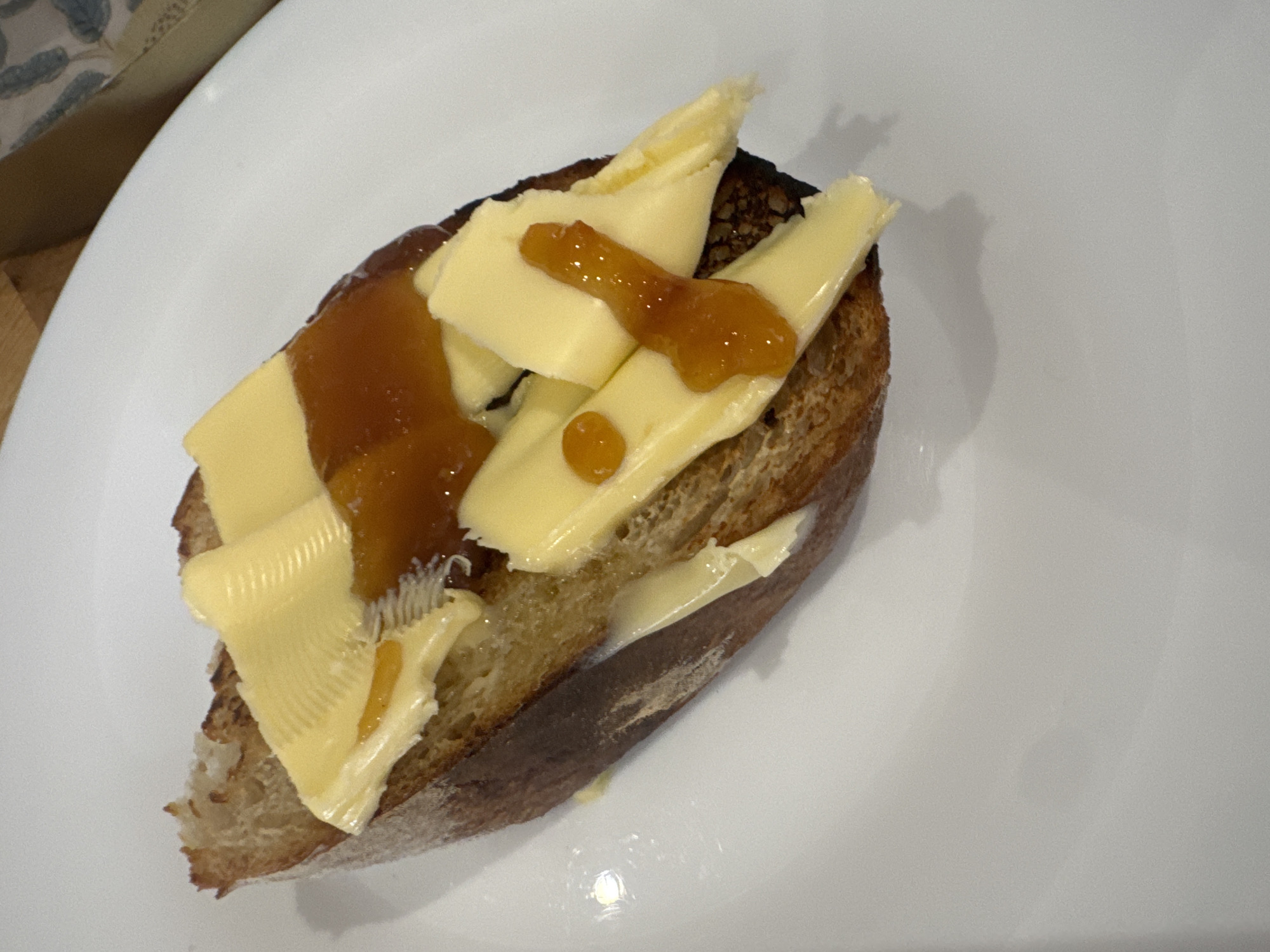 A slice of bread, topped with butter and jam, rests on a white plate. The bread is slightly toasted, with a golden-brown crust and a soft, white interior. The butter is spread in thin, wavy slices, and the jam is a rich, amber color. The plate is clean and simple, with no other objects or decorations. The lighting is soft and natural, casting a gentle shadow on the plate. There is no visible indication of the specific location in Dacorum, United Kingdom.