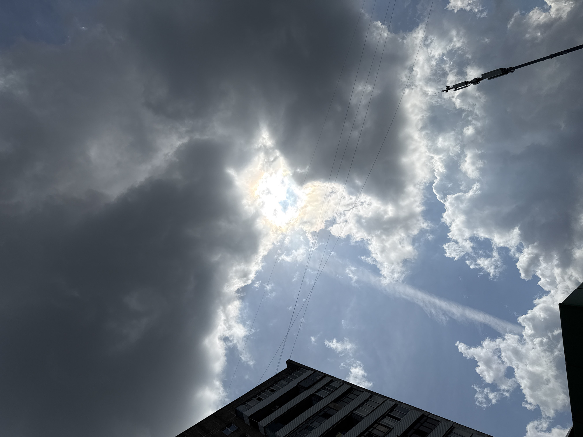 The image shows a view of the sky above a building in what is likely Moscow, Russia, although no specific location can be confirmed. Dominating the composition are various cloud formations, ranging from dark, ominous storm clouds on the left to lighter, more scattered clouds on the right. The sun is partially obscured, but its presence is evident through a bright patch in the center. Several power lines traverse the scene diagonally. In the lower portion of the frame, the top corner of a multi-story building is visible, indicating an urban environment. A pole is located on the right side of the image.