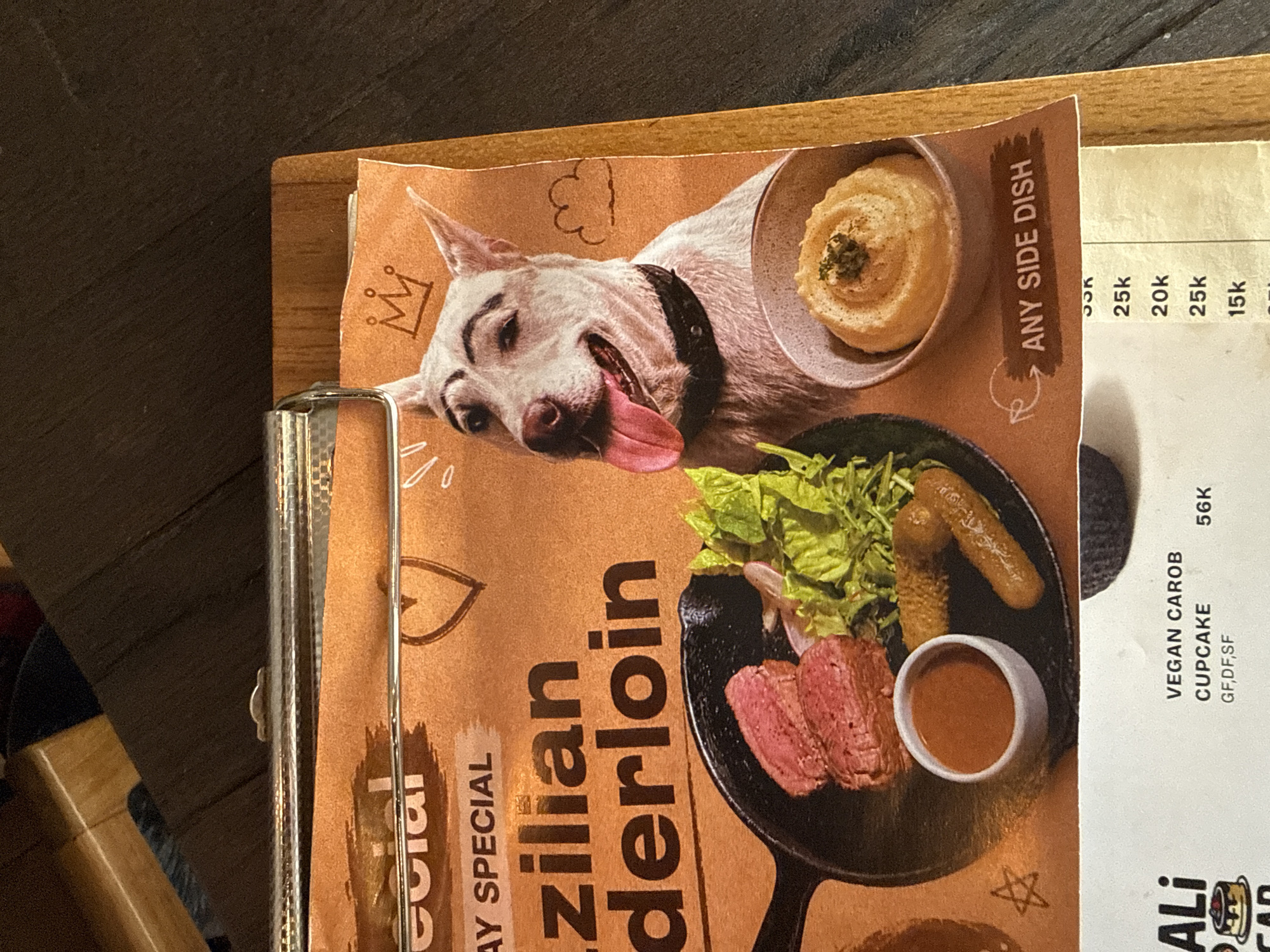 The image shows a close-up view of a menu from a restaurant, likely in Mengwi, Indonesia, as indicated by context. The menu features a humorous illustration of a dog with drawn-on eyebrows wearing a crown, placed over a picture of a Brazilian tenderloin steak served in a cast-iron skillet with a side of mashed potatoes and salad. Below the main menu item, a section lists "ANY SIDE DISH" with prices. At the bottom of the image, a portion of another menu page is visible, showing items like "VEGAN CAROB CUPCAKE" and their prices. The menu is held by a clip on a wooden board, resting on a dark wooden surface.