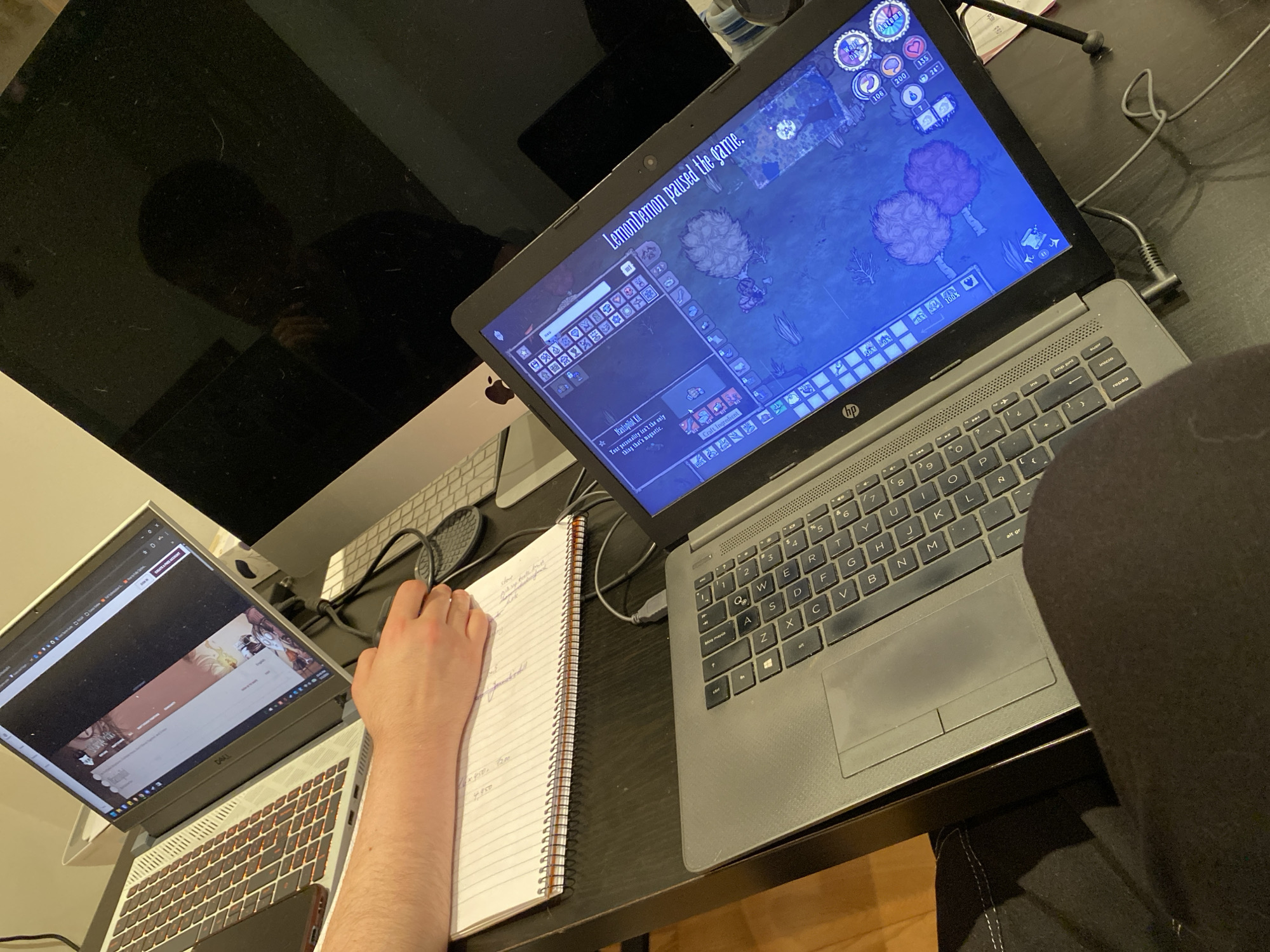 The image captures an overhead view of a multi-screen computer setup in an indoor setting, likely a home office or gaming station, located in Villaviciosa, Spain.
The primary subject is a person, whose right arm and hand are visible, resting on a spiral-bound notebook and actively writing. The rest of the person is largely obscured by the camera's perspective and a dark chair/clothing in the foreground, though a faint reflection of their head and shoulders is visible on the large, dark monitor to the left.
The desk holds three computer displays: a large, dark-screened Apple iMac monitor on the left, a silver Dell laptop to its right, and a grey HP laptop further to the right. The Dell laptop is open, displaying what appears to be a dark-themed streaming platform or social media page with some content partially visible. Underneath the Dell laptop, a smartphone is partially visible. The HP laptop is open and prominently displays a video game, "Don't Starve Together," which is currently paused. A message "LemonDemon paused the game." is clearly visible, along with game UI elements showing character stats (health 106, hunger 200, sanity 135) and the season as "Autumn." A crafting menu with items like a "Nautopilot Kit" and a description "Your personality isn't the only thing that's magnetic." is also open.
Notable details include the clutter of cables behind the laptops, connecting them to power or peripherals. The HP laptop's keyboard has a QWERTY layout with keys like "repág" and "imp pant" visible. The Dell laptop features a backlit keyboard. The notebook contains handwritten notes, including numbers like "x 450," "7200," and "4950." The overall lighting suggests an artificial indoor environment, potentially evening or night.