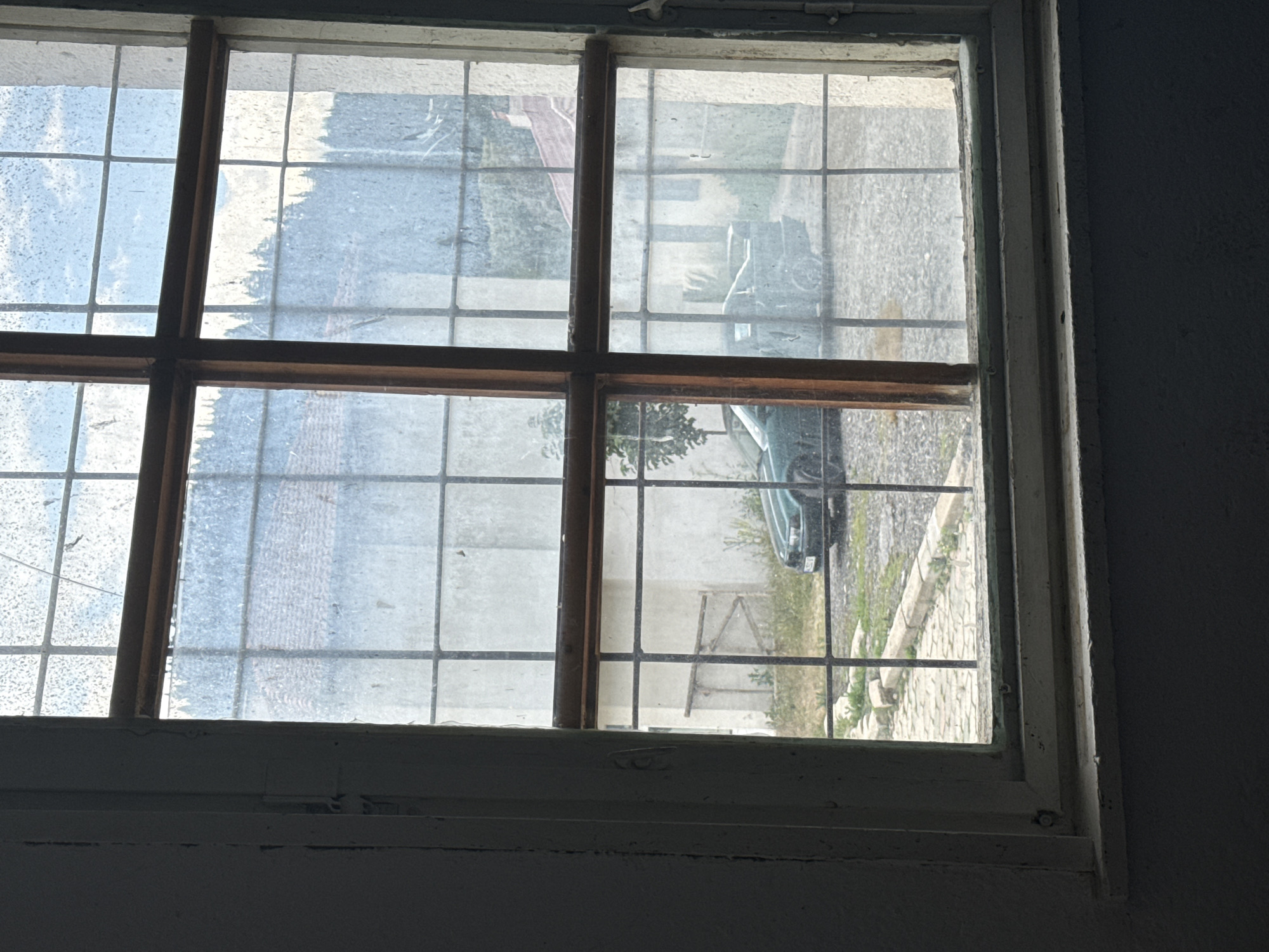 The image shows a view through a grimy, multi-paned window, likely from an interior perspective looking outward. The window frame is wooden and appears weathered. The glass is dirty and streaked, with some condensation visible. Outside, a paved area with cobblestones is visible, bordered by a concrete curb. A dark green car is parked on the paved surface. In the background, there are buildings with red-tiled roofs and a forested hill. The overall impression is one of a rural or semi-rural setting, possibly in Barutin, Bulgaria, given the context. The lighting suggests it is daytime, possibly overcast or late afternoon. No people are visible in the scene.