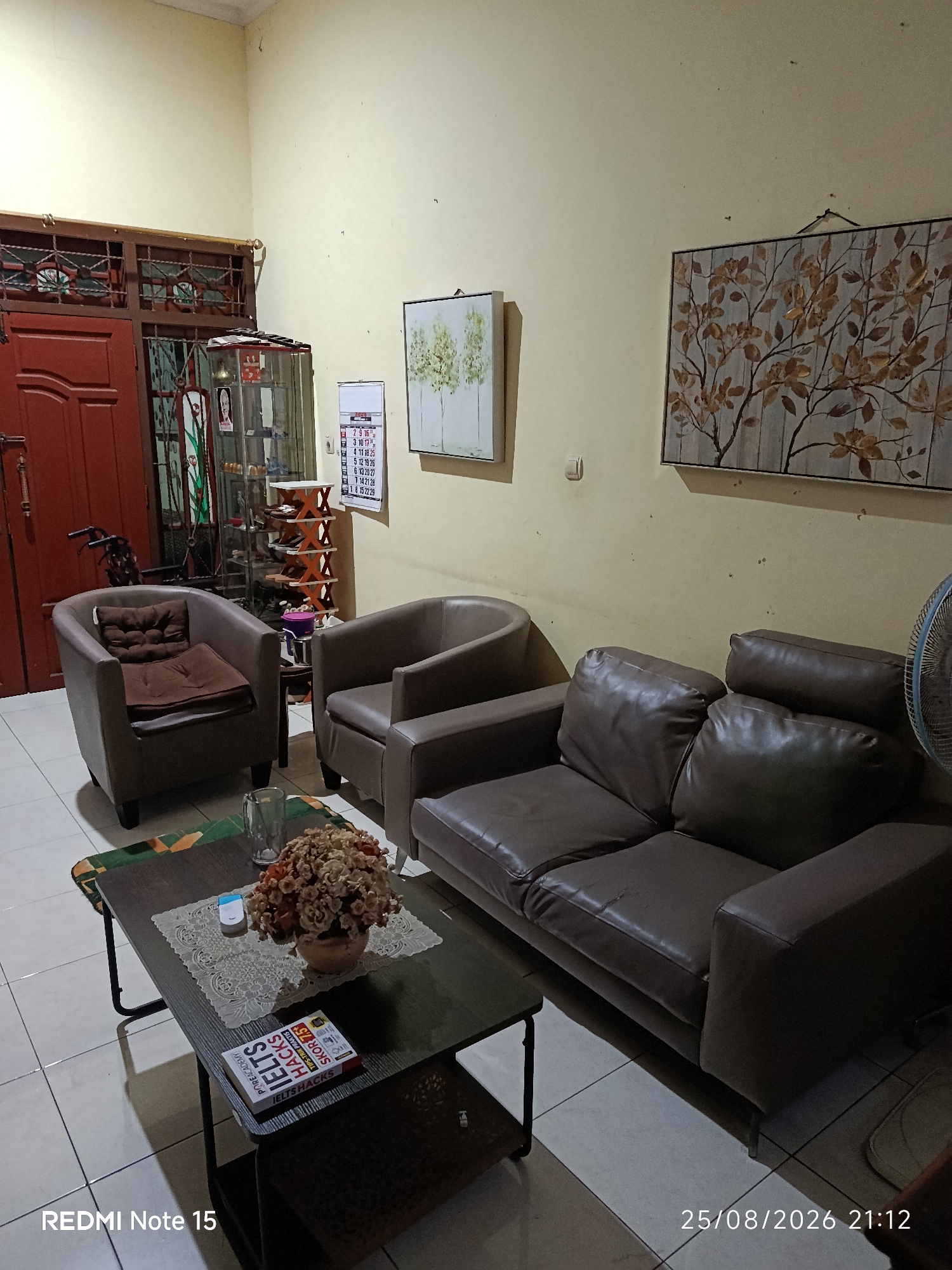In a living room, a brown leather sofa and two armchairs are arranged around a dark wooden coffee table. A glass of water, a small white electronic device, and a vase of dried flowers are on the coffee table. A book titled "IELTS Hacks" is also on the table. A calendar hangs on the wall behind the armchairs, and a framed artwork of trees hangs on the opposite wall. A wooden door with intricate metalwork is visible on the left side of the image. The room has tiled flooring and yellow walls. The timestamp "25/08/2026 21:12" and "REDMI Note 15" are overlaid on the image. The scene is indoors, suggesting a domestic setting within a city like Keboananom, Indonesia, likely a residential interior.