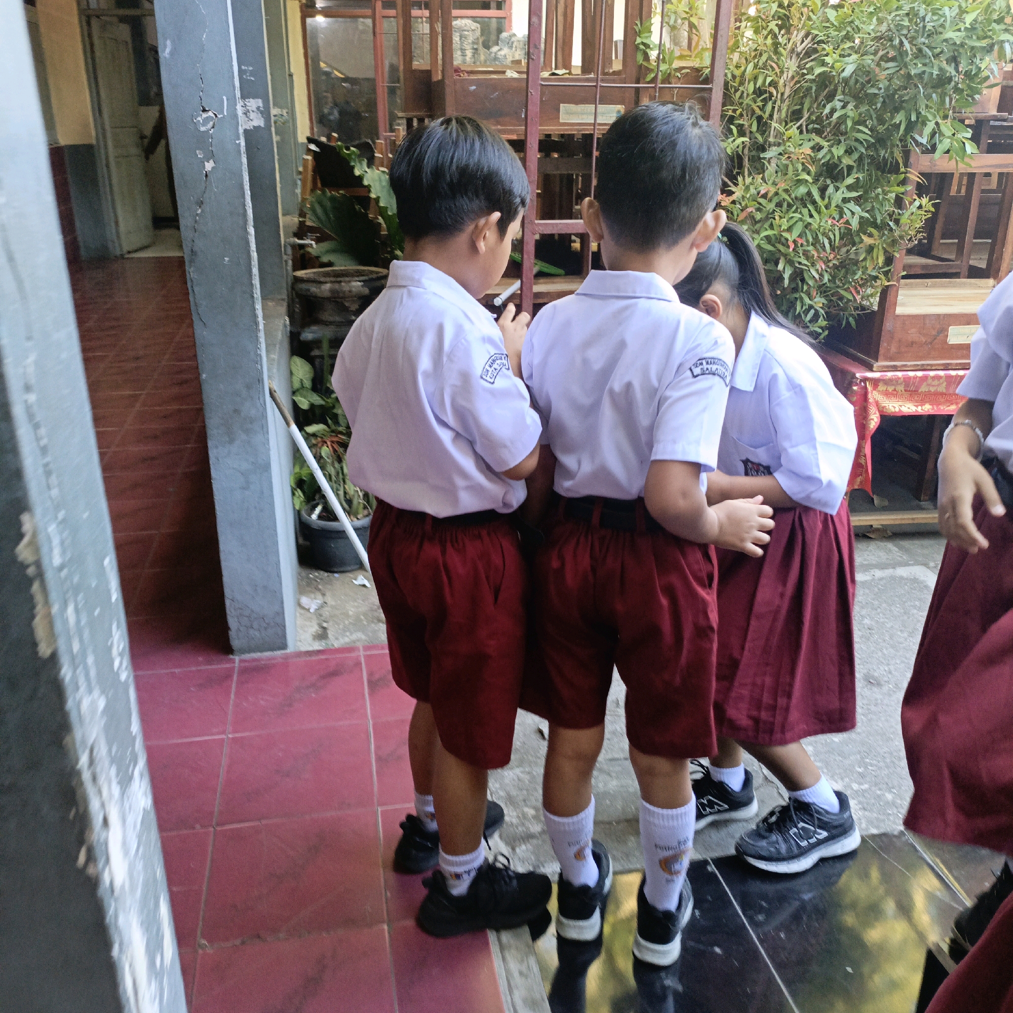 The image depicts three children, two boys and one girl, standing in what appears to be a school courtyard in Salatiga, Indonesia. The children are all wearing school uniforms consisting of white shirts and dark red shorts for the boys, and a dark red skirt for the girl. The boy on the far left is wearing a white shirt with a blue emblem on the sleeve and dark red shorts. The boy in the middle is also wearing a white shirt and dark red shorts. The girl on the right is wearing a white shirt and a dark red skirt. All three children are wearing white socks and black athletic shoes. The background shows a tiled floor, a concrete pillar with a crack, some plants in pots, and wooden structures that may be part of a school building or furniture. The lighting suggests it is daytime, with sunlight casting shadows. The overall scene conveys a typical day at an Indonesian school.