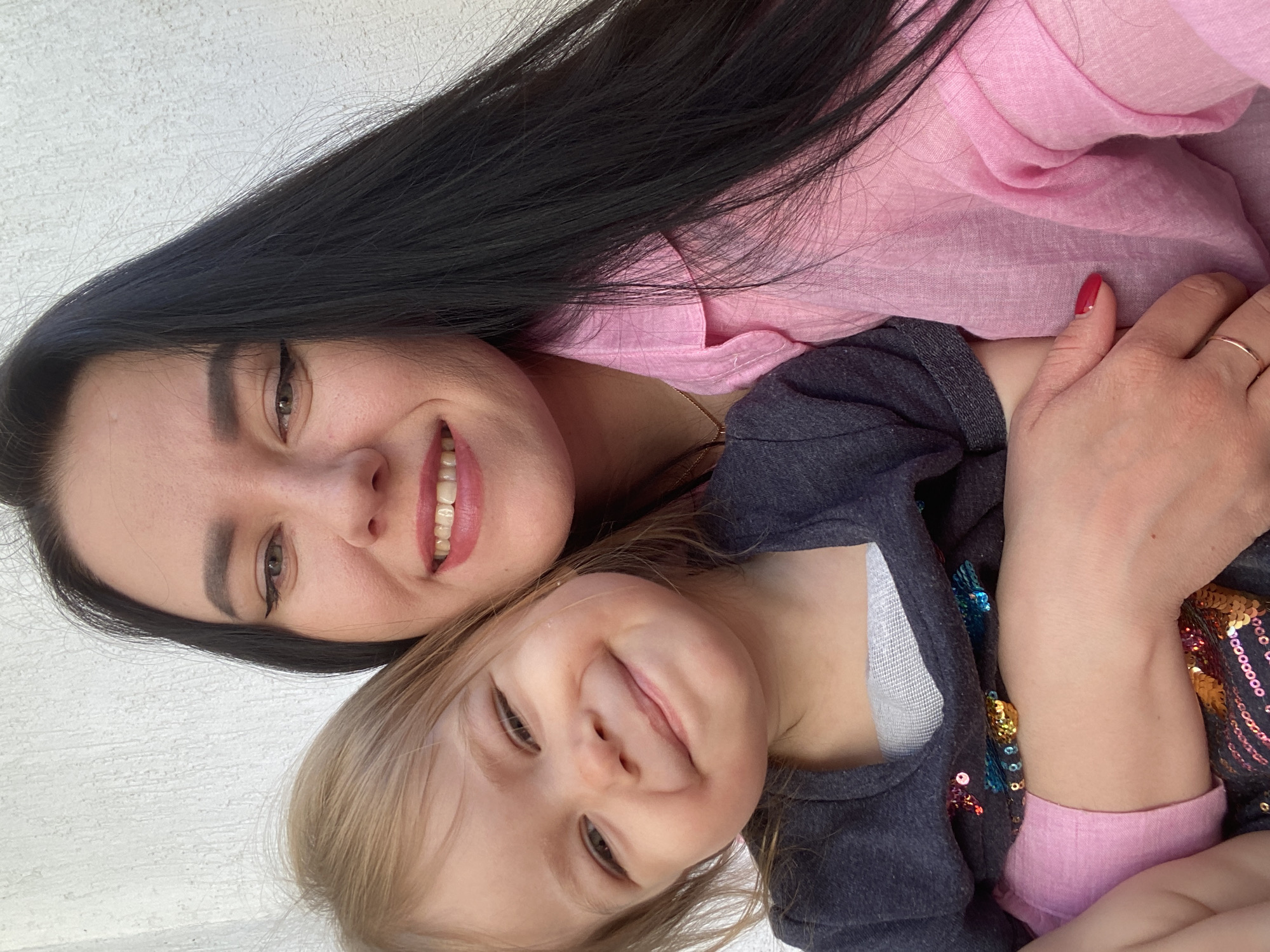 A woman with long black hair and a child with blond hair pose for a selfie in Chaikovsky, Russia. The woman wears a pink shirt, red nail polish, and a gold ring. The child wears a gray long-sleeved shirt with sequins and a white bandage on their chest. Both are smiling. The background appears to be a white or light-colored wall.
