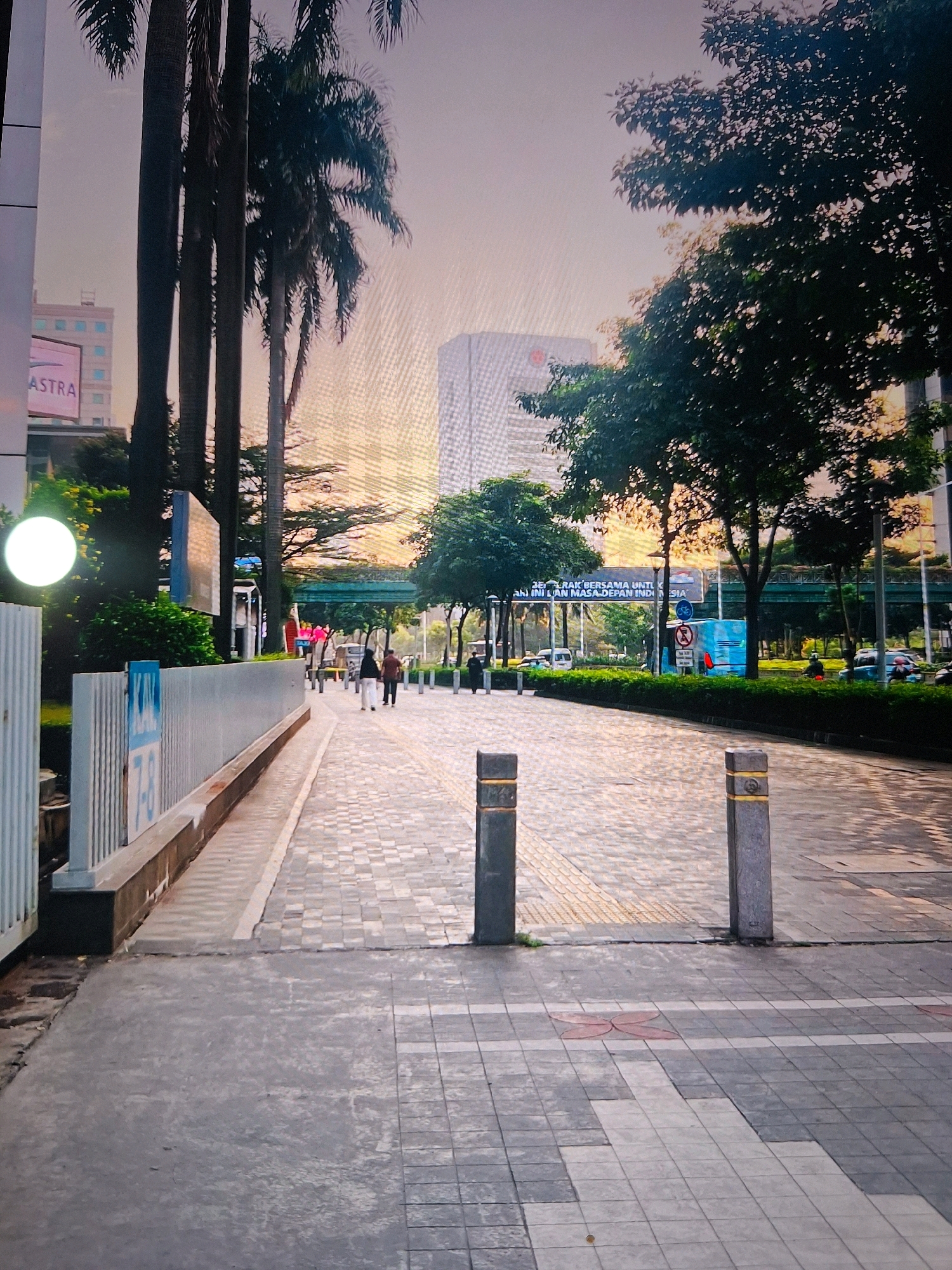The image depicts an outdoor walkway in Setiamulya, Indonesia, featuring a modern urban environment with elements of nature. Tall palm trees line the left side of the walkway, with their fronds reaching towards a hazy, sunset-toned sky. Adjacent to the palm trees is a building with a large sign that reads "ASTRA". The walkway itself is paved with interlocking gray and beige square tiles, with a distinct textured pattern for accessibility near the curb. Two gray bollards with yellow bands are positioned at the entrance to the main pedestrian area.
On the right side of the walkway, lush green bushes form a neat hedge. Beyond the hedge, several trees with dense foliage are visible. In the background, modern buildings rise above the trees, and a pedestrian bridge spans across the street. The sky is bathed in a soft orange and pink hue, suggesting it is either sunrise or sunset.
Several people are seen walking along the paved walkway. Two individuals are walking away from the camera towards the left, wearing dark clothing. Further down the walkway, more people are visible in the distance. The presence of vehicles, including cars and a blue truck, suggests this is a busy area. A large billboard with text in Indonesian is visible in the background, above the pedestrian bridge. The overall atmosphere is that of a well-maintained urban public space.