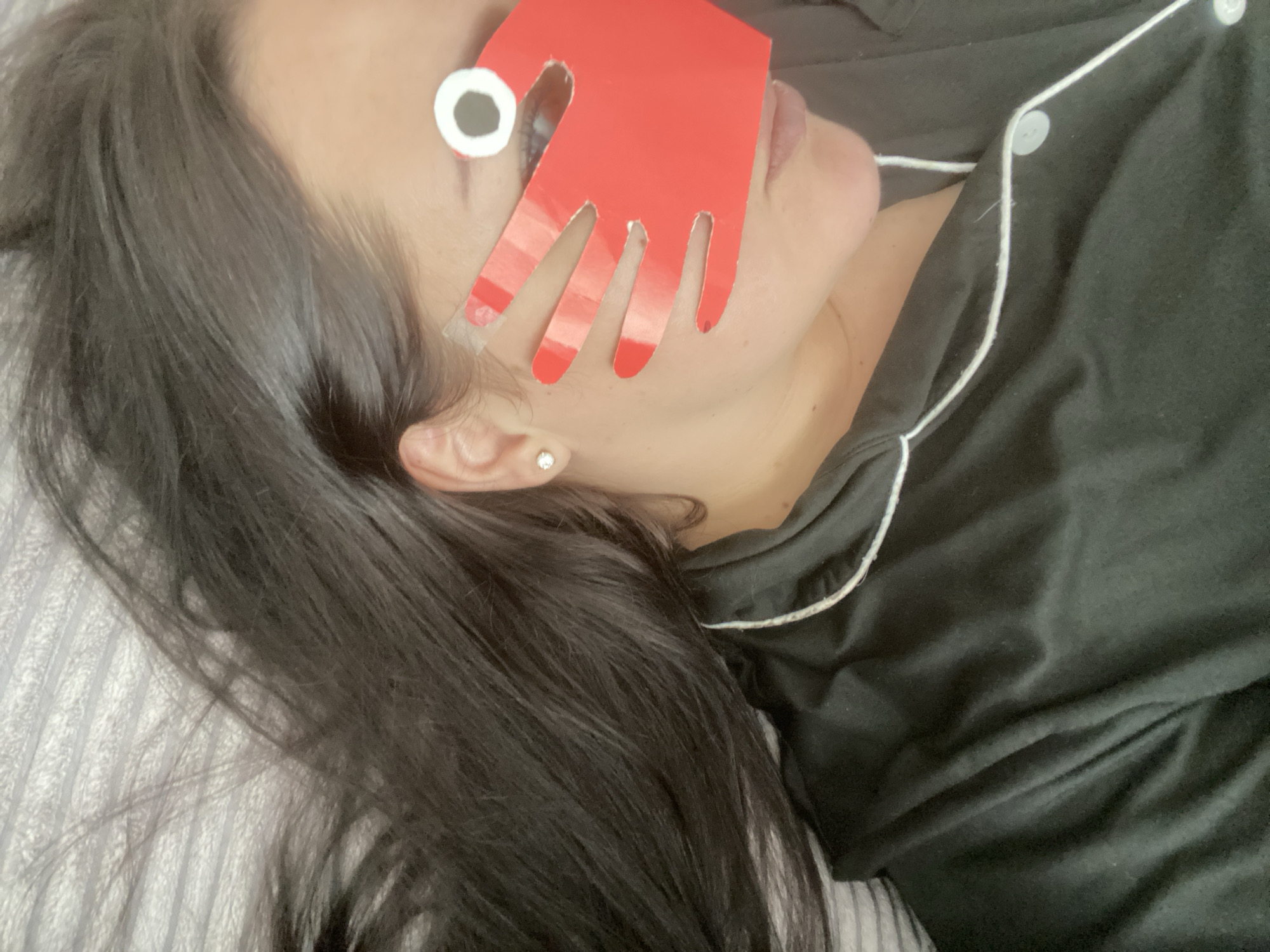 A woman lies in bed, obscured by a red, hand-shaped cutout with a white circle attached to the index finger. She is wearing black pajamas with white piping. The background is a striped blanket or sheet. No information in the image suggests this is in Sochi, Russia.