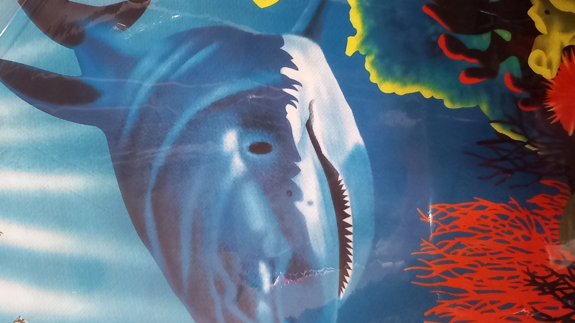 The image is a close-up, dynamic, and colorful illustration of a shark in an underwater scene, likely part of a child's toy or decorative item. The shark, rendered in shades of blue with white accents, dominates the frame, its mouth agape revealing sharp, white teeth. Its eye is a dark oval, and its body shows stylized patterns, suggesting movement or water currents.
The background depicts a vibrant coral reef. Bright yellow and green formations create an impression of sunlight filtering through the water. Red and dark coral structures add depth and visual interest to the scene. The overall aesthetic is cartoonish and engaging, aiming to capture the imagination.
There are no people visible in the image, nor any specific activity or event taking place beyond the depiction of the shark in its natural habitat. The setting is clearly an ocean environment, characterized by the shark and coral. There are no discernible cues about the time of day or weather, as it's an artistic representation. The emotional tone is one of excitement and perhaps a touch of awe associated with marine life.
Visible text is minimal and appears to be a small, white logo or inscription in the lower left portion of the image, but it is too indistinct to be read. The location context of Kos, Greece, does not directly inform the content of the image itself, as the imagery is generic to an underwater theme.