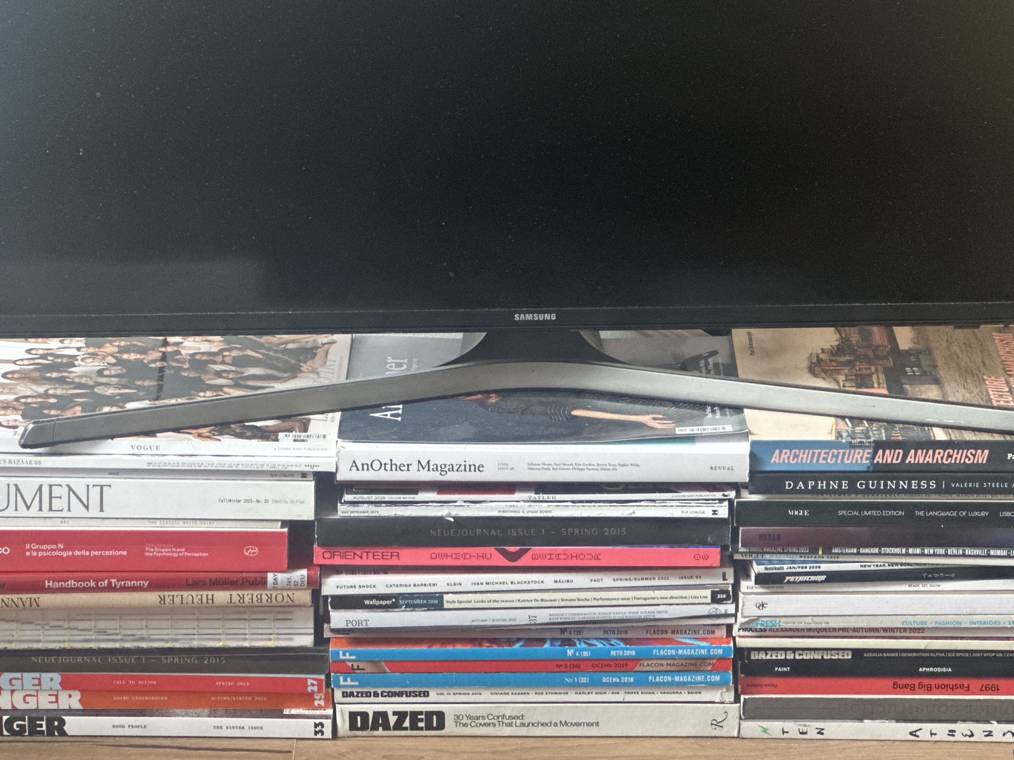 A stack of magazines supports a Samsung monitor. The titles of the magazines include "Vogue," "Harper's Bazaar," "AnOther Magazine," "Architecture and Anarchism," "Daphne Guinness," "Document," "II Gruppo N," "Handbook of Tyranny," "Norbert Heuler," "Neuejournal Issue 1 - Spring 2015," "Ger Ger," "Dazed & Confused," "Orienteer," "Tatler," "Wallpaper," "Port," "Flacon Magazine," and "Men's Health." The magazines are stacked in rows, with the monitor's base resting on top. The location is not specified.
