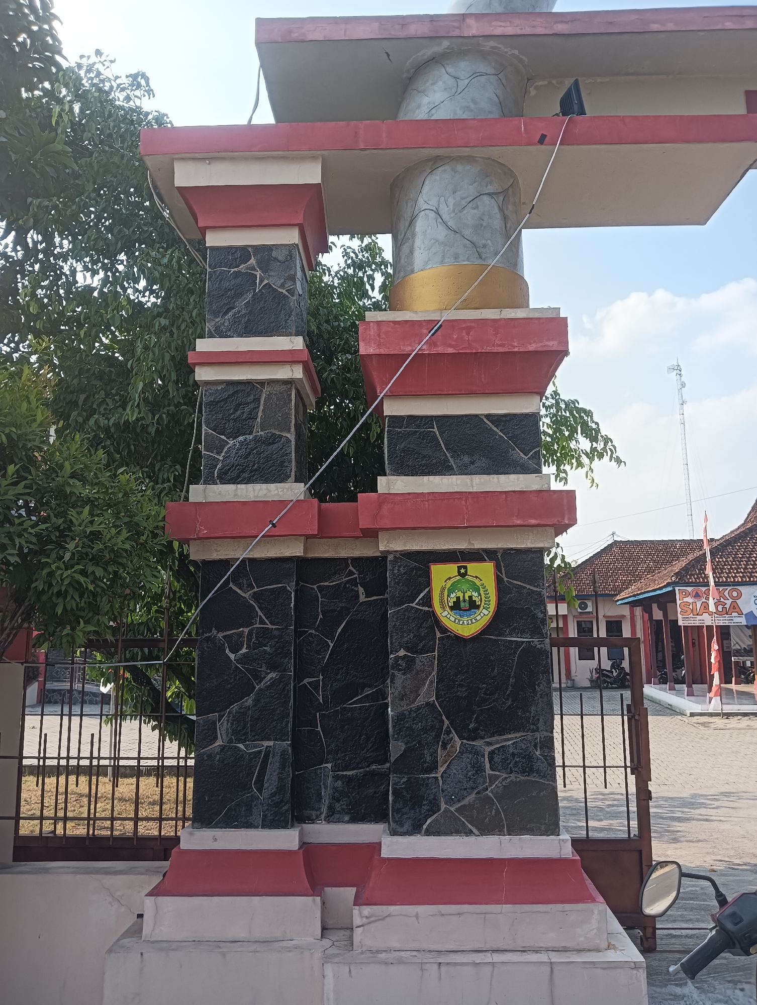 This image captures a portion of an entryway structure located in Palemrejo, Indonesia. The structure is made of concrete and stone, with prominent red and white accents. Two large, stone-clad pillars form the entrance, topped by red and white rectangular frames. A gold band encircles a cylindrical element on the structure. A yellow and green shield emblem is affixed to the left pillar. Behind the structure, a building with red-tiled roofs is visible, along with trees, a metal fence, and a paved area. A motorcycle is partially visible in the lower right corner. The scene is set under a clear sky with some scattered clouds.