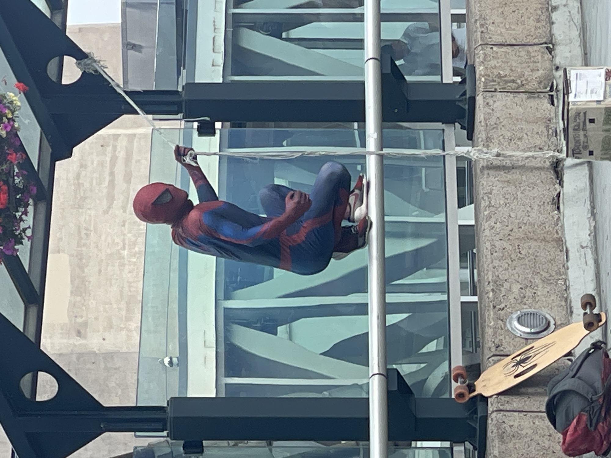 An individual dressed as Spider-Man is crouching on a horizontal railing, holding a strand of what appears to be white string. The Spider-Man costume is red and blue with red webbing details. The person is wearing athletic shoes. Behind them, a large building with a glass facade is visible. Above the Spider-Man, a dark metal structure with a circular opening and hanging flower baskets is present. In the foreground, a longboard with a spider design is leaning against a concrete barrier, next to a red and black backpack. Another person, out of focus, is visible in the background wearing a light-colored shirt. The overall scene appears to be an outdoor urban environment.