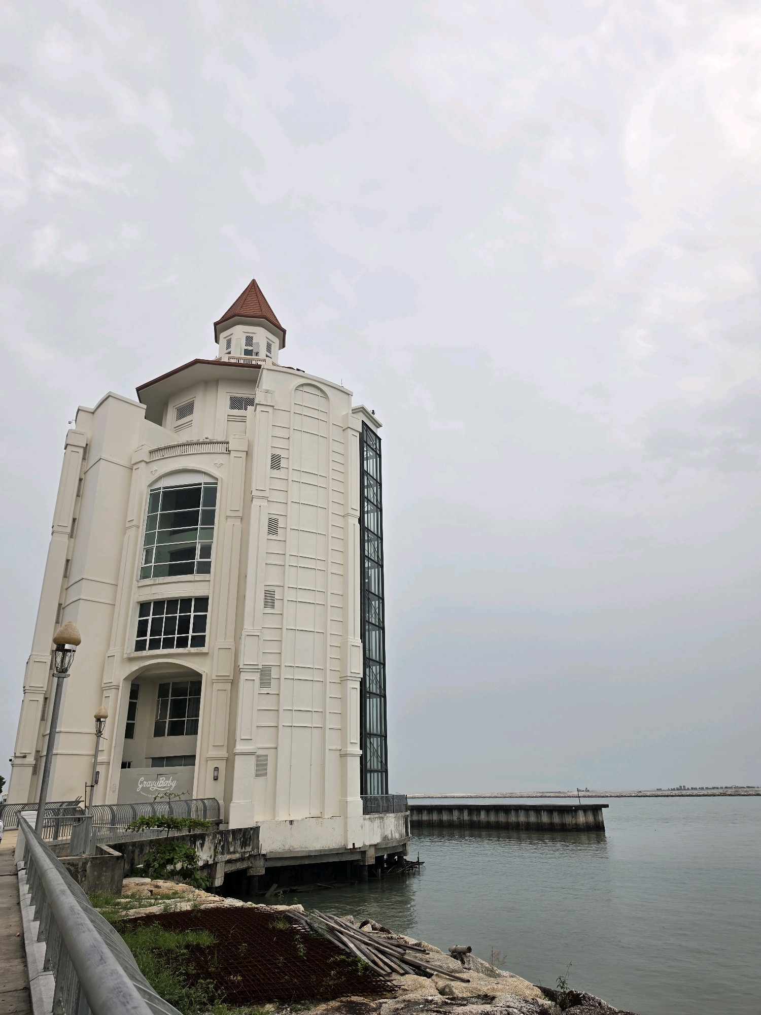A tall, white, multi-story building with a conical red roof stands on the water's edge in George Town, Malaysia. The building has large windows, an exterior elevator, and ornate architectural details. The water is calm, and a pier extends into the distance. The sky is overcast.