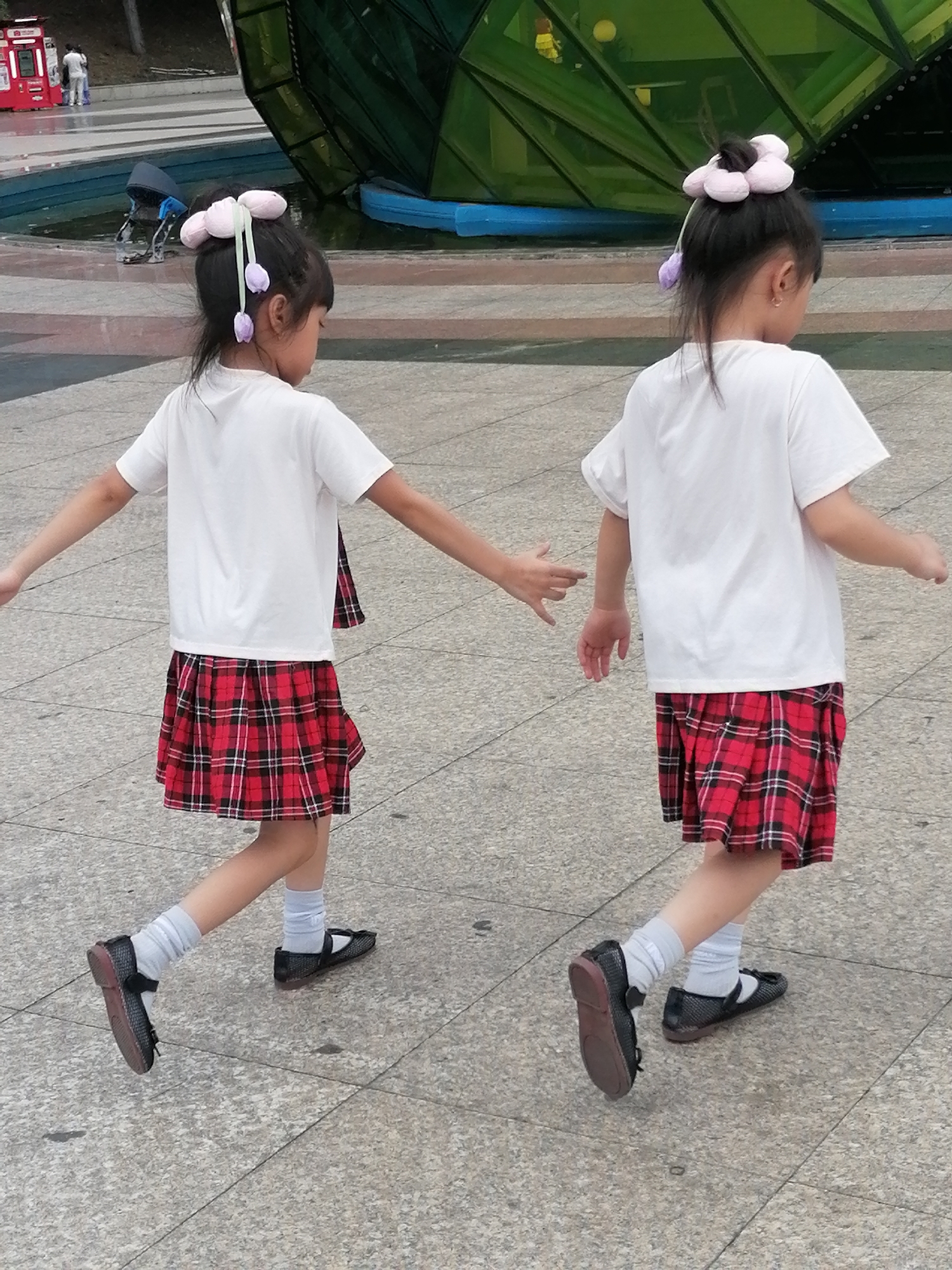 Two young girls, each with distinctive pink flower-shaped hair accessories and purple tulip-shaped dangling charms, are walking away from the camera on a paved outdoor surface in what appears to be a public area. They are both wearing white short-sleeved shirts, red and black plaid skirts, white socks, and black flat shoes. The girl on the left has her left arm extended slightly outwards, while the girl on the right has both arms down by her sides. Behind them, a large green, multifaceted structure is visible, possibly an architectural element or sculpture, with a blue edge along its base. In the background, there is a body of water with a blue pool liner and a few people standing near a red vending machine. The lighting suggests an overcast day. The scene is consistent with a location in Da Lat, Vietnam, known for its pleasant climate and public recreational spaces.
