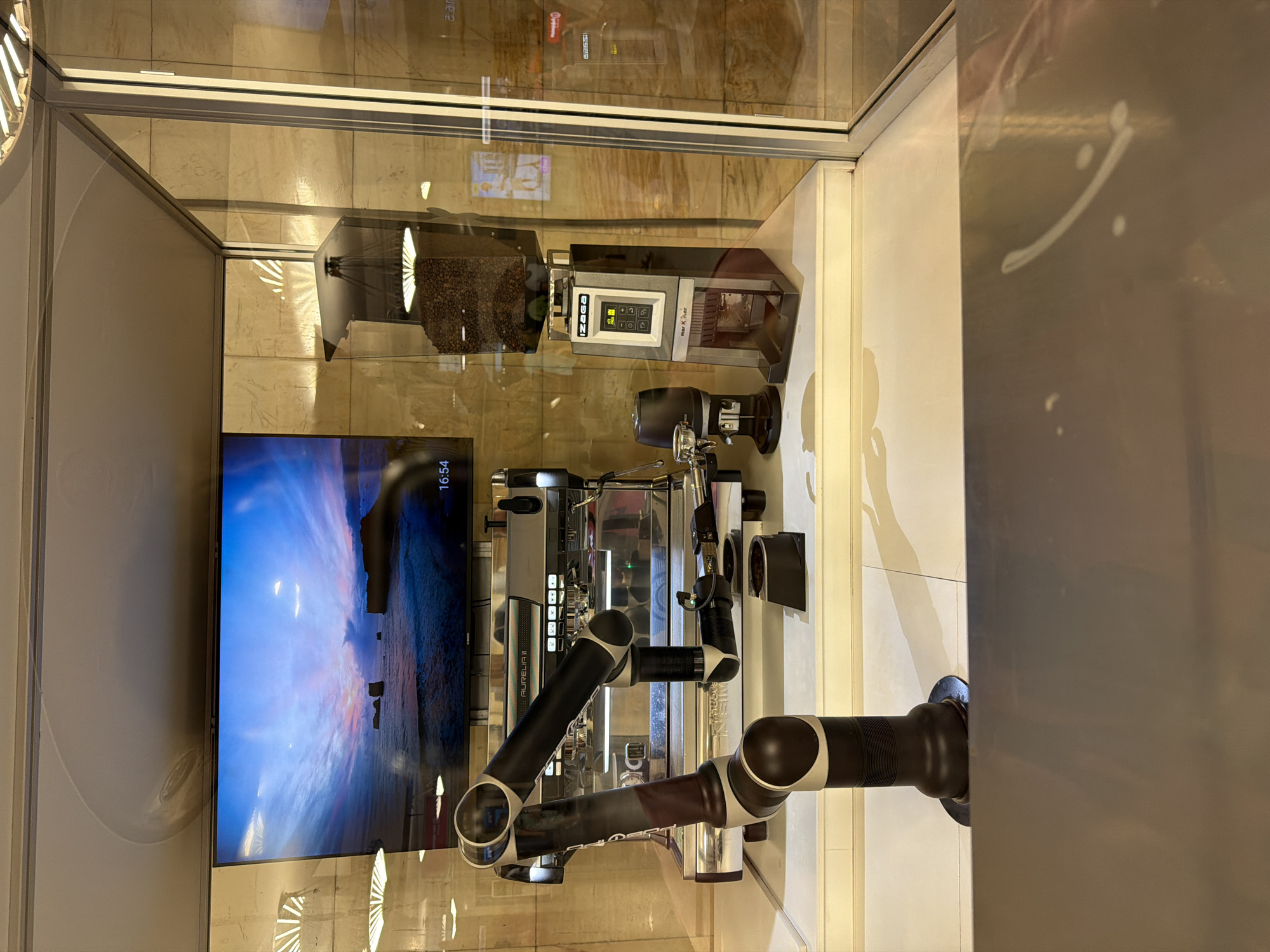 The image shows a robotic coffee machine in a glass display case. Behind the coffee machine is a television screen displaying a landscape scene. Above the coffee machine is a coffee bean grinder. The location is Moscow, Russia.