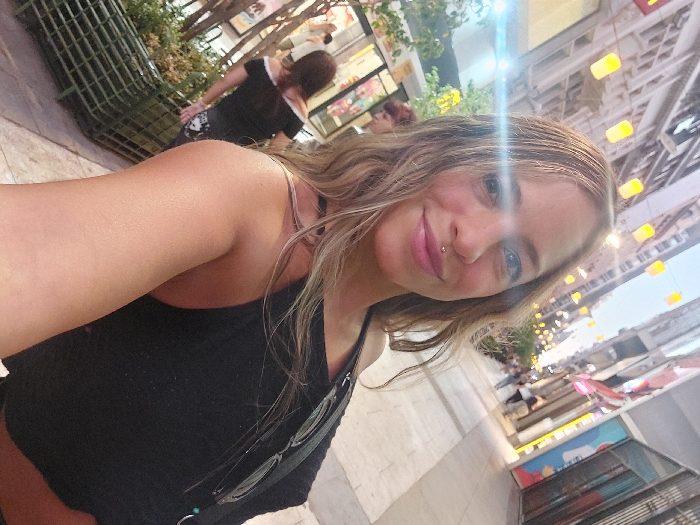 A fair-skinned woman smiles at the camera in a selfie. She has blonde hair and a labret piercing. She wears a black tank top and has sunglasses hanging from the neckline. The background is an outdoor scene with blurred figures and buildings. Lanterns are strung overhead. There is a green bench and plants visible in the scene.