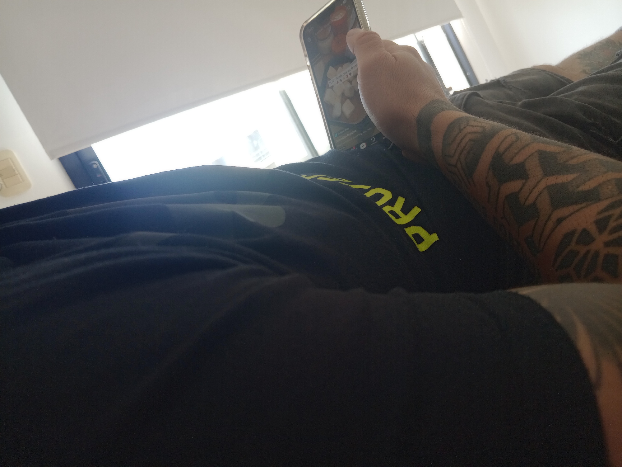 A person is lying down, holding a smartphone in their hand. The smartphone is displaying a video, possibly from TikTok, showing food being prepared. The person has tattoos on their forearm, including a large geometric design. They are wearing a dark-colored garment with the word "PRU" visible in yellow lettering. The setting is indoors, next to a window with daylight coming through. The location is identified as Lomas de Zamora, Argentina.