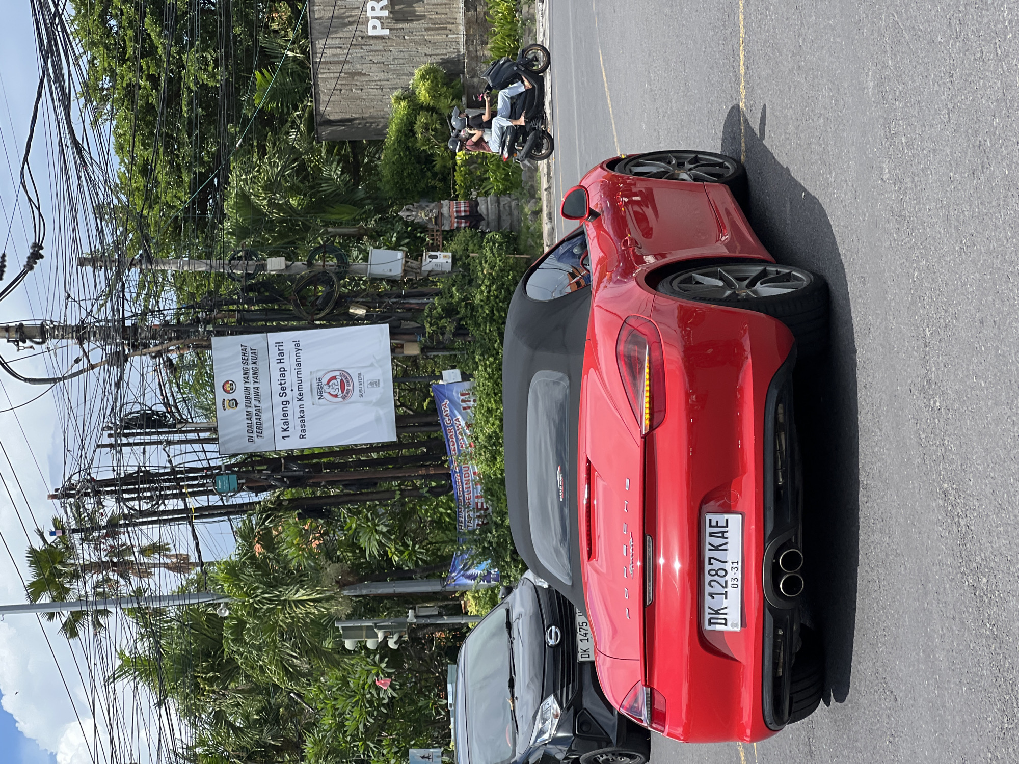 The image captures a vibrant street scene, likely in Denpasar, Indonesia, characterized by a mix of vehicles and dense urban infrastructure under bright daylight.
In the foreground, a glossy red convertible sports car, identifiable as a Porsche Boxster by its rear badging, is parked or stopped on the side of a paved road. It displays the Indonesian license plate "DK 1287 KAE," with an expiration date of "03-31." Directly in front of it is a black van or SUV, also with an Indonesian license plate reading "DK 1475." Further down the road, a scooter carries two individuals, both wearing helmets, one of whom has their arm raised. Another scooter is partially visible further ahead.
The setting features numerous utility poles laden with a chaotic tangle of overhead electrical and communication wires, a common sight in some urban areas of Indonesia. Lush green foliage, including palm trees and other dense tropical vegetation, lines the street and forms a backdrop. A building with the partial text "PR" is visible on the right. A stone statue or carving is also seen amidst the greenery. The sunny weather casts strong shadows, indicating a mid-morning or afternoon time.
Several banners and signs are affixed to the utility poles. A prominent white banner reads: "DI DALAM TUBUH YANG SEHAT TERDAPAT JIWA YANG KUAT" (In a healthy body, there is a strong soul), followed by "1 Kaleng Setiap Hari! Rasakan Kemurniannya!" (1 Can Every Day! Feel the Purity!), and features the logos for "Nestle" and "BEAR BRAND SUSU STERIL" (Sterile Milk). A partially obscured blue banner also displays Indonesian text, including fragmented words like "GAYA HIDUP SEHAT" (Healthy Lifestyle) and "PELINDUNGAN" (Protection).