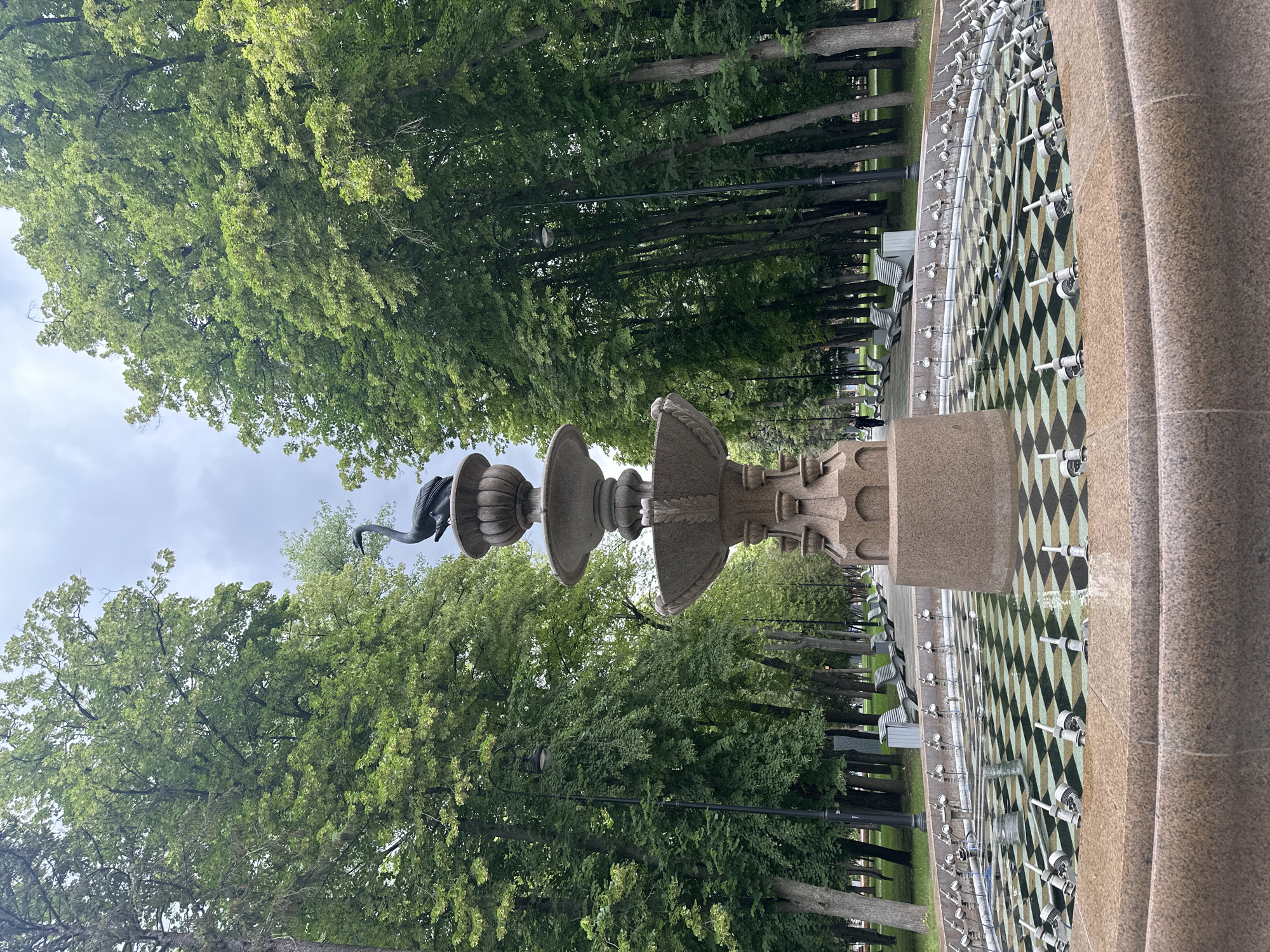 An ornate, multi-tiered stone fountain stands in the foreground of a park. The fountain features a large basin with a checkered pattern at the bottom, and tiered bowls above. At the very top, a dark, bird-shaped statue is perched.
Behind the fountain, a tree-lined path stretches into the distance, with benches visible along the sides. Streetlights are positioned at intervals along the path. A single person is walking away from the camera on the left side of the path. The sky is overcast, and the trees are full of green leaves. The overall scene depicts a serene, public park setting.