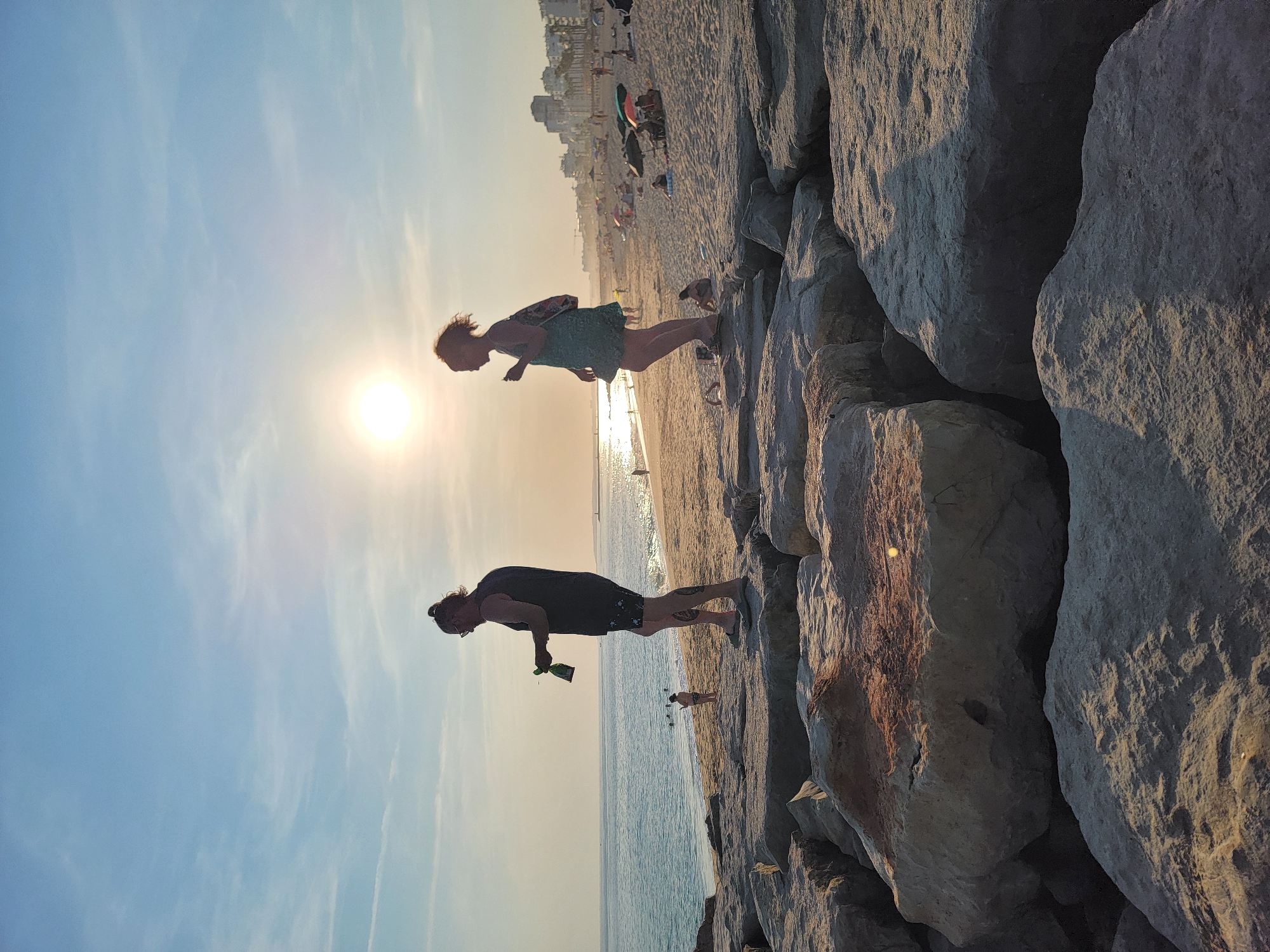 The image shows a beach scene in Quarteira, Portugal, during sunset. In the foreground, two people are standing on a rocky embankment. A man with his hair in a bun and wearing a dark tank top and shorts is holding a bottle. A woman in a light-colored dress is standing next to him. The background depicts a sandy beach with several people relaxing under umbrellas, a calm ocean, and the city skyline. The sun is low in the sky, casting a warm glow over the scene and reflecting on the water.