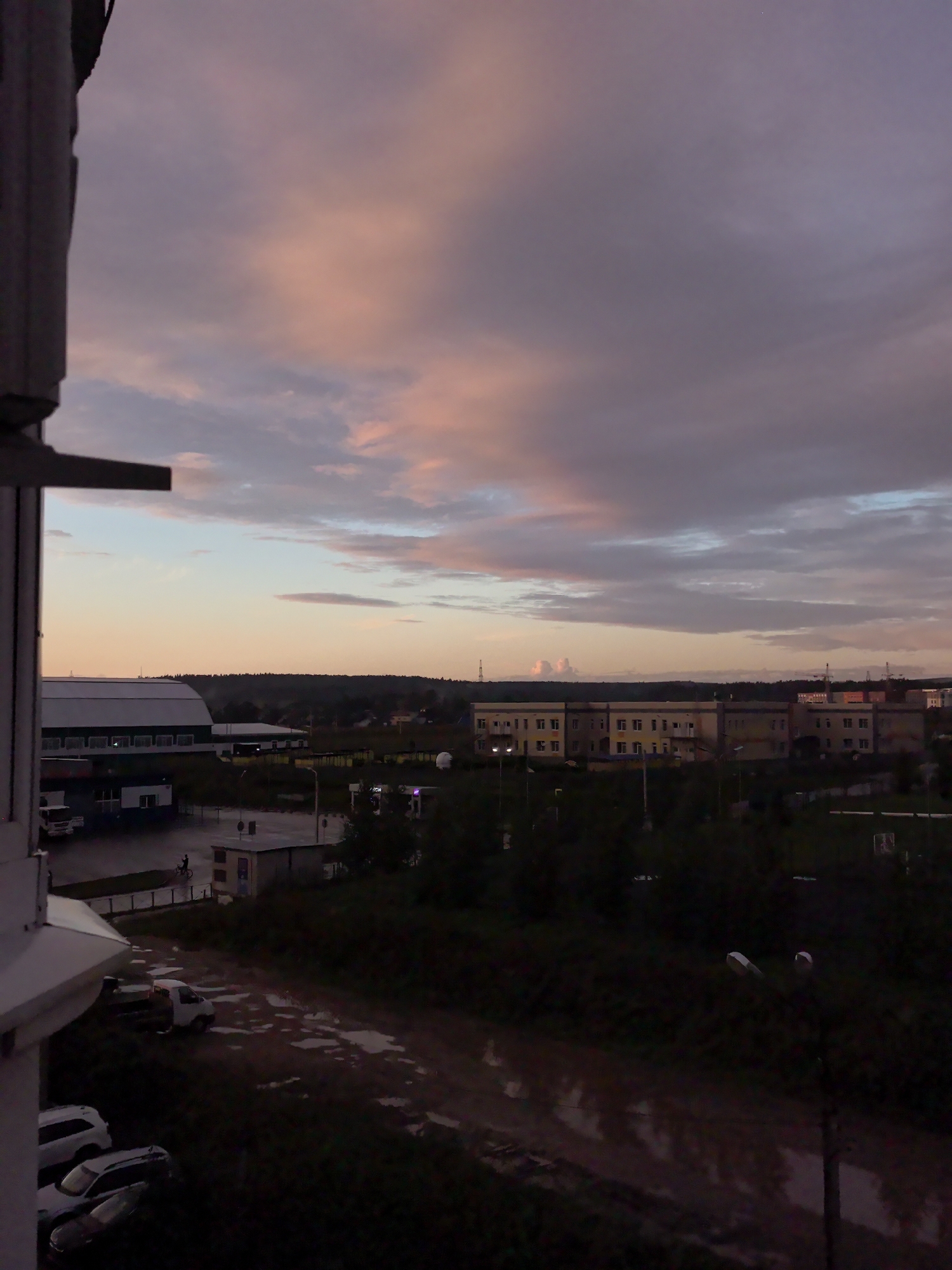 The image is a medium shot of a town in Froli, Russia, at dusk. The sky is a blend of purple, pink, and blue clouds, with the last light of the sun casting a warm glow on the horizon. In the foreground, a portion of a building's exterior wall is visible on the left. The middle ground shows a street with several vehicles, including a white truck, a white SUV, and a dark sedan. A person is riding a bicycle on the wet street, which has numerous puddles reflecting the sky. In the background, there are several low-rise buildings and a line of trees against the fading light. The overall atmosphere is subdued and peaceful, suggesting a quiet evening in a small town.