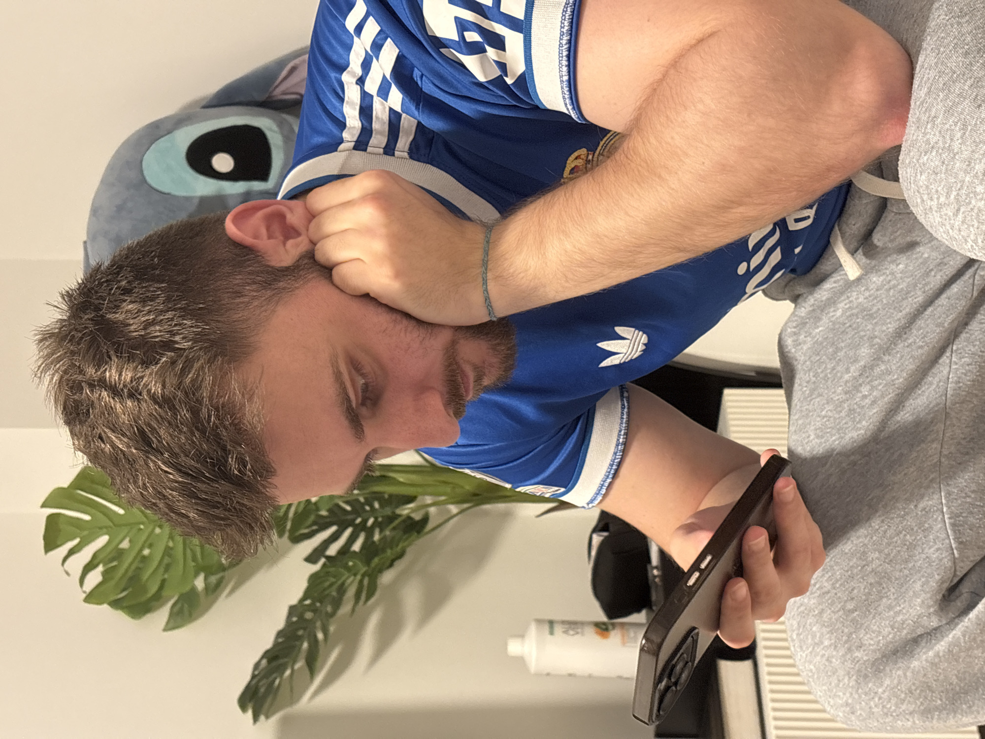 A young man with light-brown hair and a beard is sitting down and looking at a cell phone. He is wearing a blue soccer jersey with white stripes on the sleeves and gray sweatpants. His face is resting on his hand. Behind him is a stuffed animal that appears to be Stitch from the movie Lilo and Stitch. There is a green plant and a white tube of lotion behind the phone.
There is no information in the image to confirm the location is in Mönchengladbach, Germany.