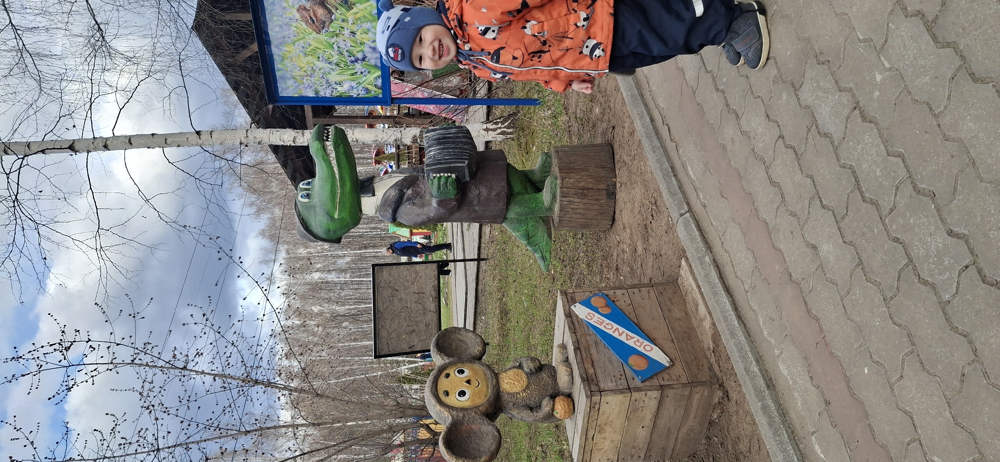The image shows a young child standing beside a wooden sculpture of Gena the Crocodile and Cheburashka in what is likely a park or recreational area. The statues are situated on a paved walkway next to a grassy area with trees. In the background, there are other trees and structures, including a sign with an image. The child is wearing a winter hat and jacket, suggesting a cool climate. The presence of the Gena the Crocodile and Cheburashka sculpture indicates the location is likely in Russia, since these characters are from Russian children's literature. The location is the city of Chaikovsky, Russia.