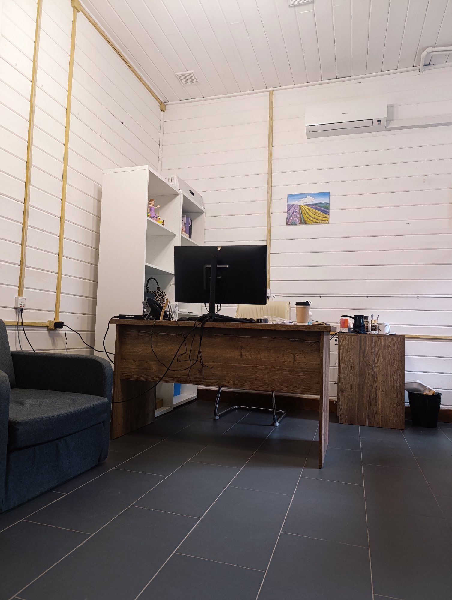 This image shows an office setting. A dark-colored armchair sits to the left. A desk with a dark wood finish is in the center, with a computer monitor positioned on top, its screen facing away from the camera. Cables are visible behind the desk. To the left of the monitor, there is a white shelving unit filled with various items. To the right of the desk, there is a small wooden cabinet, and next to it, a black trash can.
The walls are white with horizontal paneling, and vertical wooden beams run along them. On the wall to the right, there's a painting of a lavender field and an air conditioning unit is mounted above it. The floor is covered with dark gray tiles. The location is identified as Unknown, Unknown.