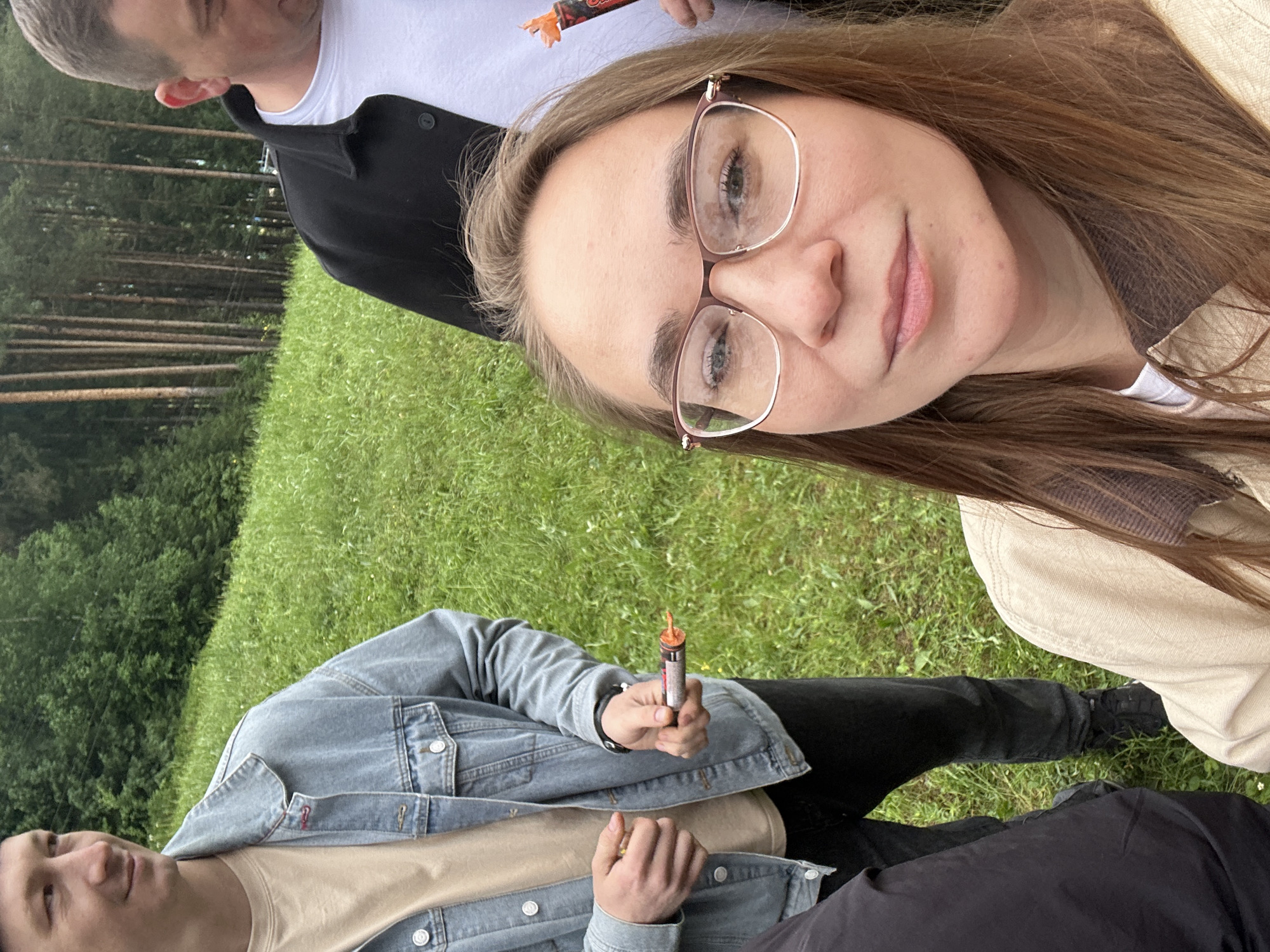 A woman with brown hair and glasses takes a selfie. Two men are visible; one wearing a black jacket and the other a jean jacket. Both men are holding cylindrical objects that are orange at the top. The setting is a grassy area with trees in the background. The location is Прикамский, Russia.