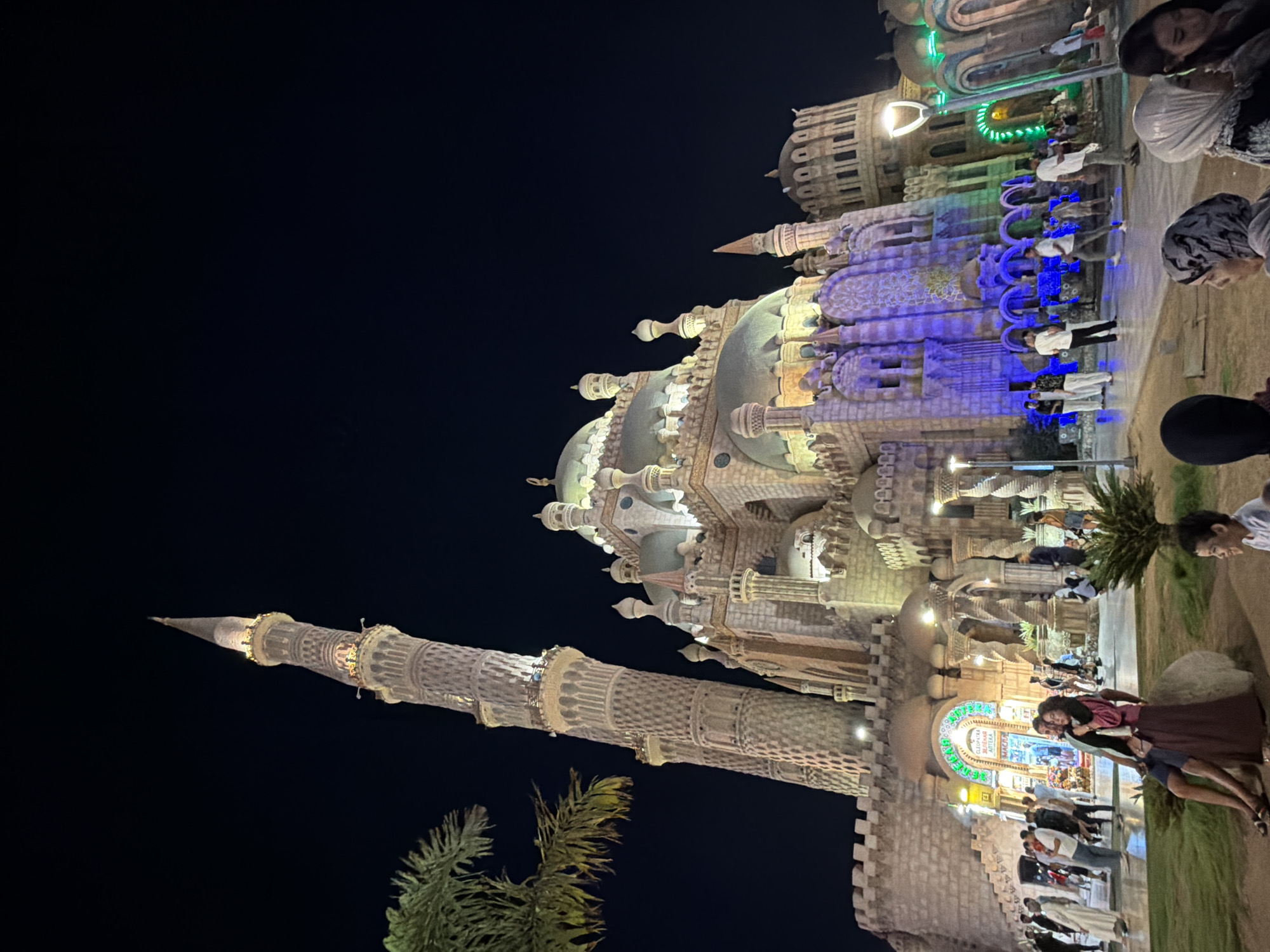 This image captures the imposing facade of the Al Sahaba Mosque at night in Sharm El Sheikh, Egypt. The mosque, with its intricate minaret and domes, is illuminated by artificial lights, casting a warm glow against the dark night sky. Palm fronds from a nearby tree frame the left side of the image, adding a touch of tropical scenery. In the foreground, several people are visible, some walking and others sitting. A couple is seated on a rock in the center of the frame, their faces turned towards each other. Other individuals are scattered throughout the scene, creating a sense of activity and life around the architectural marvel. The overall atmosphere is one of grandeur and evening serenity.