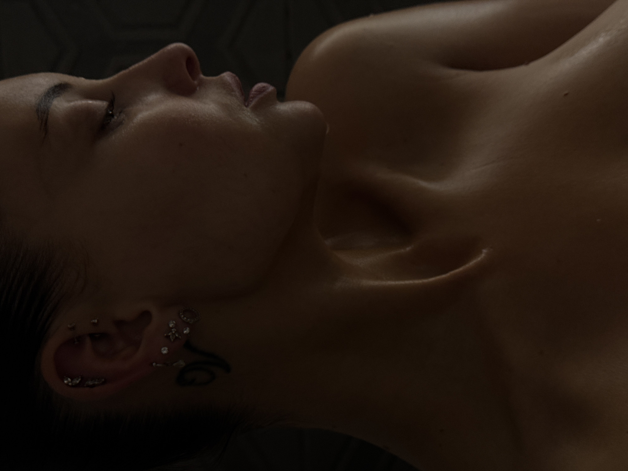 A close-up, dimly lit portrait features a young adult female lying down, viewed in profile from her left side. Her eyes are closed or nearly closed, suggesting a state of rest or sleep. Soft, dramatic lighting illuminates the contours of her face, highlighting her cheekbone, nose, and slightly parted lips. Her skin appears smooth and slightly dewy or oily, catching highlights along her jawline, neck, and collarbone.
Her dark hair is visible around her ear. Her left ear is adorned with numerous piercings, including star-shaped studs and various small, decorative jewels distributed across the helix, conch, and tragus areas. A dark, abstract tattoo, possibly a swirling or tribal pattern, is visible on her neck below her ear.
Her upper body is bare, revealing her collarbone and the gentle curve of her shoulder and décolletage. The background is uniformly dark and out of focus, creating a subdued and intimate atmosphere. The overall impression is one of peacefulness and serenity, captured in a private, indoor setting, possibly a bedroom or a spa environment. There is no visible text in the image.