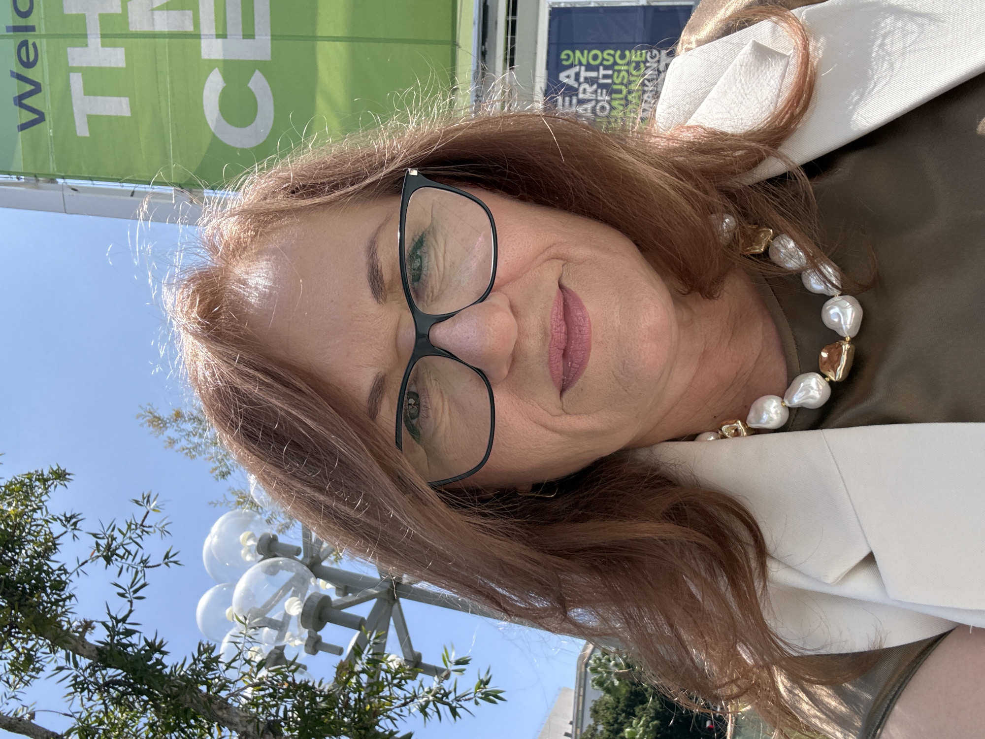 A medium shot captures a woman with auburn hair and glasses, looking directly at the camera. She is outdoors, with a bright blue sky and green foliage in the background. A large green banner with white text that reads "THE M C" is visible to her right, suggesting the location is a venue or establishment. To her left, a streetlight with multiple globes is partially visible, and further left, there's a hint of a building. The woman is wearing a light-colored blazer over a dark olive-green top, and a distinctive chunky pearl necklace with gold accents. The overall impression is a sunny day in an urban or park-like setting within Los Angeles.
