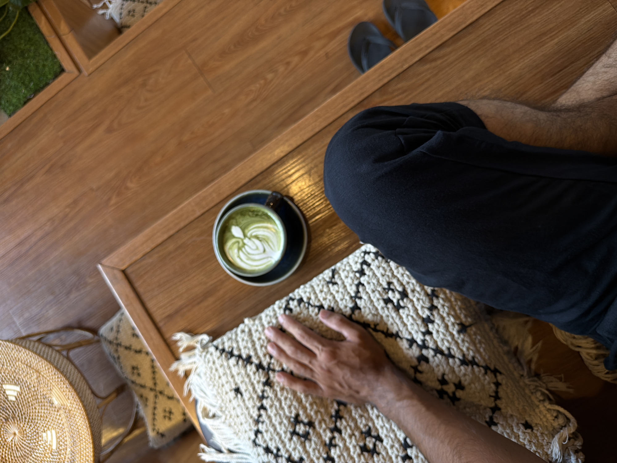 A high-angle shot depicts a person sitting at a wooden table in what appears to be a cafe or indoor lounge, possibly in North Kuta, Indonesia. The person is wearing dark-colored clothing and flip-flops; their right hand rests on a textured, cream-colored rug with a geometric black pattern and fringe. A cup of green beverage, likely matcha latte, sits on a dark saucer on the table next to the rug. The table is made of brown wood with noticeable grain. To the left are hints of a woven basket and a decorative cushion. In the background, a shelf-like structure containing greenery can be observed.