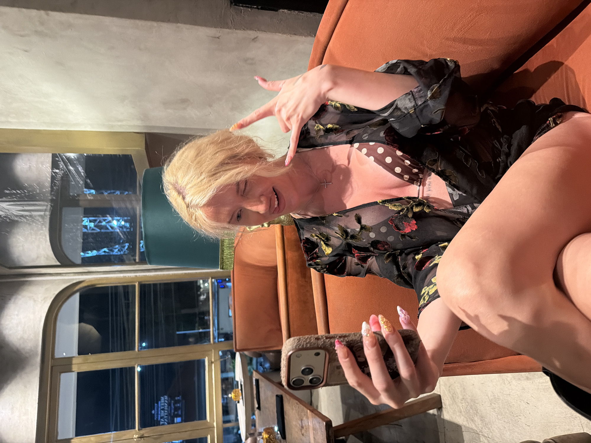 A blonde woman is seated on an orange couch in a dimly lit establishment, possibly a bar or restaurant, in North Kuta, Indonesia. She is holding a smartphone in her left hand, taking a selfie, and winks at the camera while sticking out her tongue. Her right hand is raised, forming a "rock on" gesture. She is wearing a sheer black floral-print robe over a polka-dot bikini top. The background shows a large window with city lights visible outside, and a teal lampshade. A wooden table with some items on it is also visible to the left of the woman.