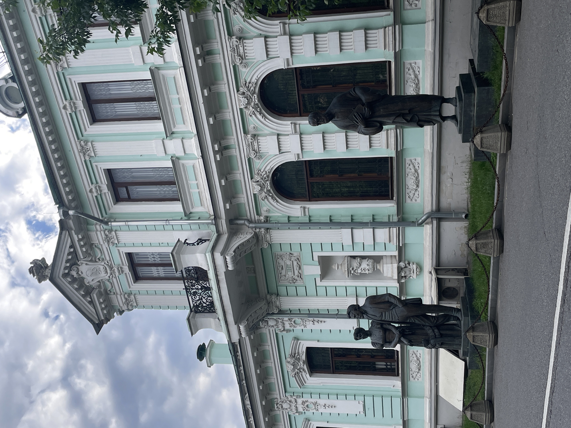 The image shows the exterior of a pale green building with white architectural details, featuring arched windows and ornate stonework. In front of the building, there are three bronze statues of people, positioned on a raised platform. The statues appear to be of historical figures, with one man and two women depicted. They are dressed in formal attire, and their stances suggest they are in conversation or observation. The building's facade exhibits classical architectural elements, including columns, pediments, and decorative carvings. A black wrought-iron balcony is visible on the upper floor.
To the right of the statues, a paved road runs alongside a strip of green grass, separated by a low barrier with decorative stone planters and a metal chain. In the background, the sky is partly cloudy, suggesting daytime. The overall scene appears to be an outdoor public space, possibly a park or square adjacent to a significant building in Moscow, Russia. No visible text is present in the image.