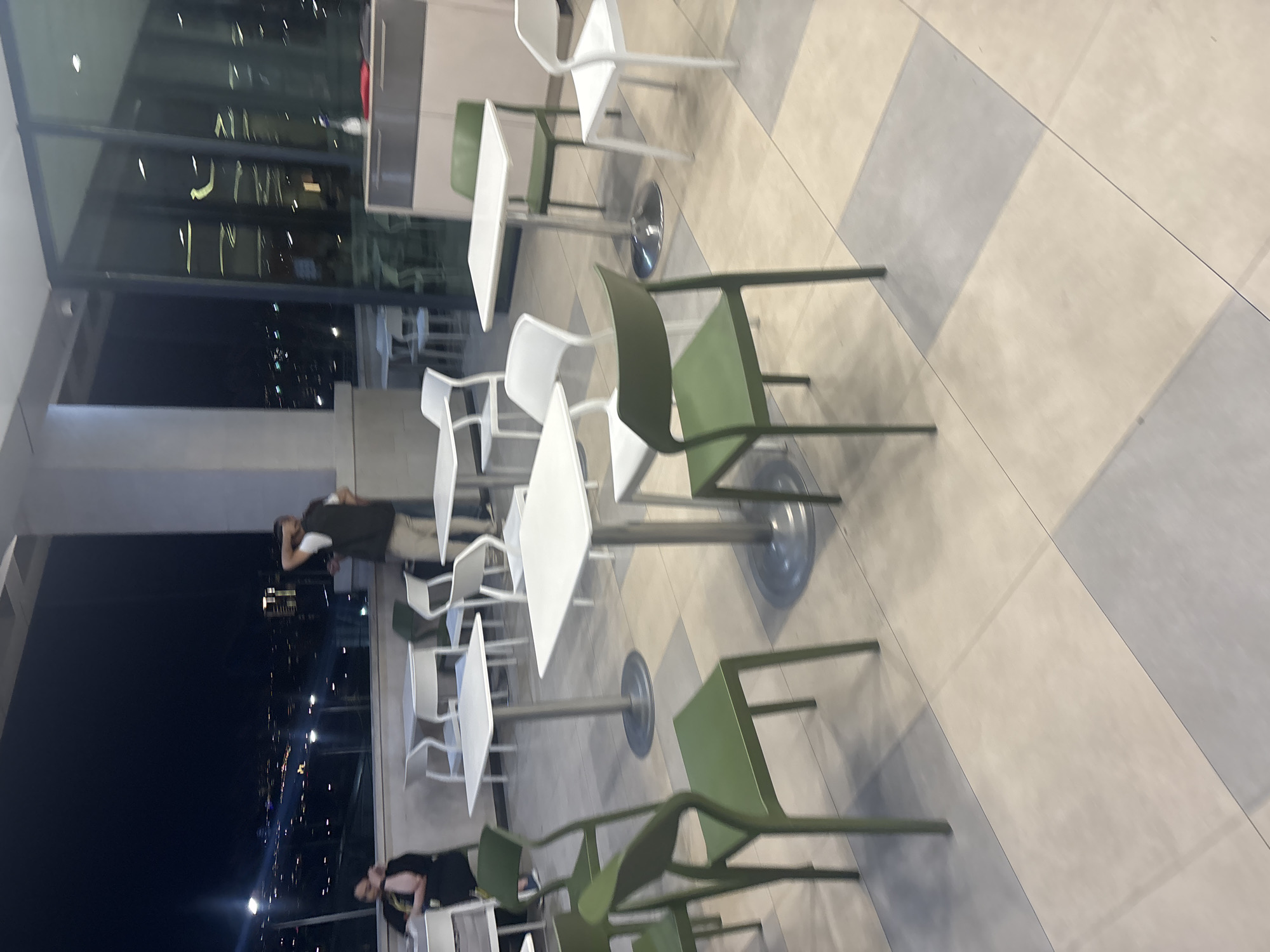 The image, initially oriented vertically but best viewed horizontally (rotated 90 degrees counter-clockwise), depicts a modern outdoor or semi-outdoor dining area at night, overlooking a city in Yerevan, Armenia.
The scene features several square white tables with chrome bases, paired with a combination of white and olive green chairs. These are arranged on a checkered floor with alternating light and dark grey tiles. A large glass barrier separates this seating area from the exterior view.
Through the glass, a dark night sky is visible, illuminated by countless distant city lights, suggesting a high vantage point overlooking an urban landscape. Reflections of the interior lighting and furniture are faintly visible on the glass surface.
Two individuals are present. On the left side of the frame, a person wearing a dark top and light pants stands, appearing to lean against a wall or railing while looking out at the city lights. Another person is partially visible in the bottom right, seated at one of the tables.
The overall atmosphere suggests a relaxed, evening setting, possibly a rooftop restaurant or lounge. The image has a slightly soft focus, particularly on the background elements. No visible text is present.