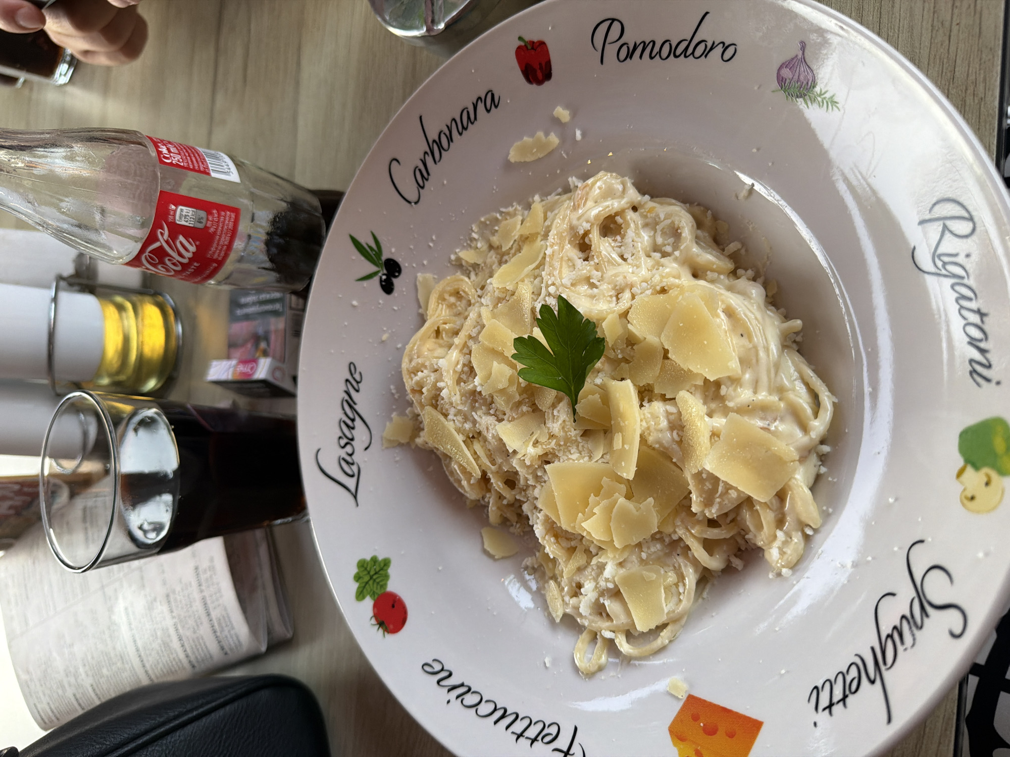 The image shows a plate of pasta, likely fettuccine or spaghetti carbonara, served with a creamy sauce and topped with shaved parmesan cheese and a sprig of parsley. The plate is decorated with various pasta names and illustrations, including "Fettuccine," "Lasagne," "Carbonara," "Pomodoro," and "Spaghetti."
On the table, there is a glass of dark liquid, possibly cola, next to a partially consumed bottle of Coca-Cola. A pack of cigarettes is also visible. In the background, a menu or newspaper is partially obscured by a black handbag.
The setting appears to be an outdoor cafe or restaurant with a wooden table. The overall lighting suggests it is daytime. The location is identified as Svishtov, Bulgaria.