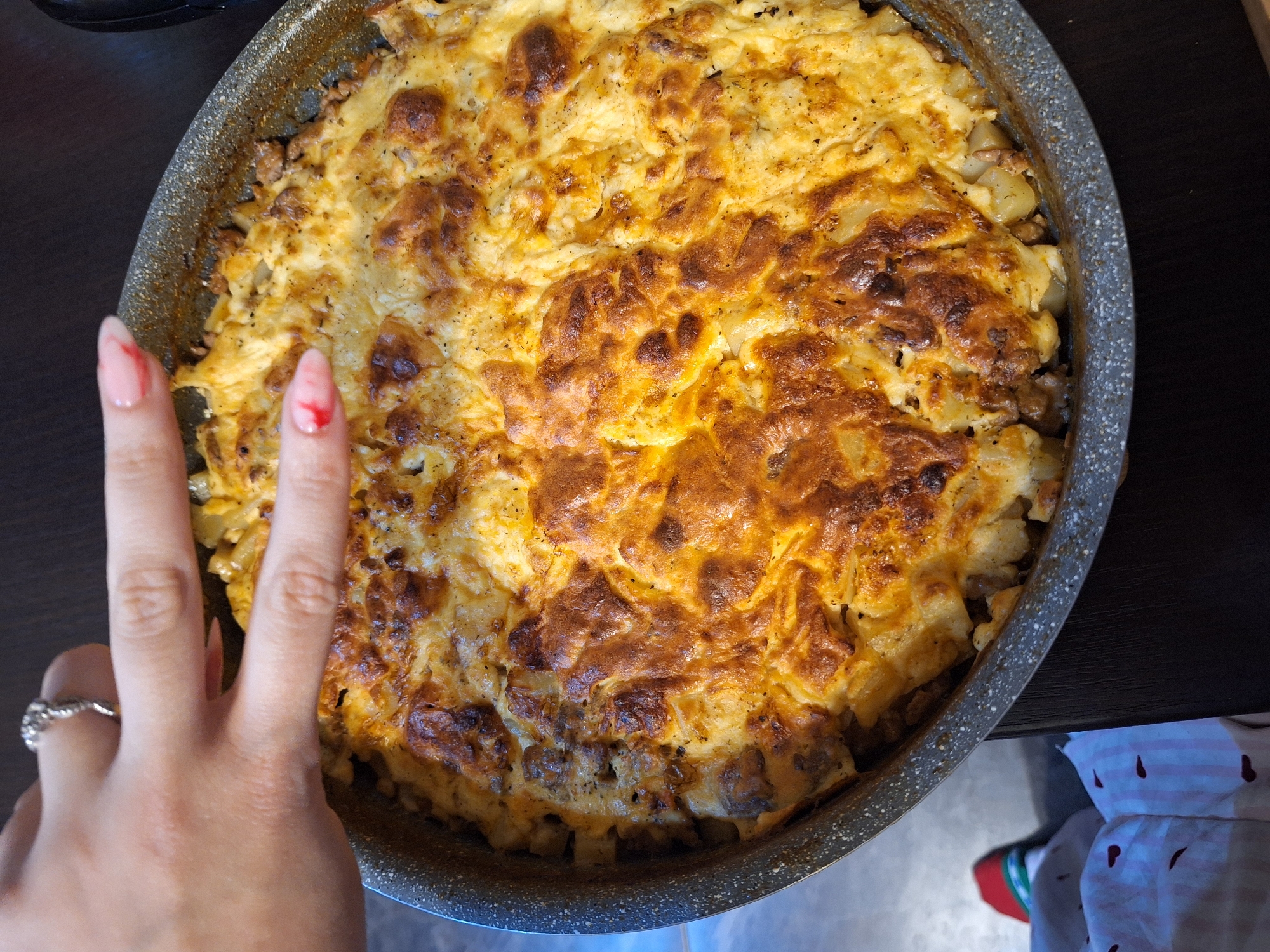 A high-angle shot displays a baked dish, possibly a casserole or gratin, in a dark grey pan. The dish has a golden-brown, somewhat bubbly crust, suggesting it has been baked in an oven. Visible underneath the crust, there appear to be cubed potatoes and possibly meat or another vegetable.
To the left of the pan, a person's hand is visible, with two fingers extended in a peace sign gesture. The person has a ring on their finger and their nails are painted light pink with a red decorative pattern on one finger.
The pan is placed on a dark, smooth surface, likely a table or countertop. In the lower right corner, the edge of a patterned fabric (possibly an apron or tablecloth) with red heart motifs is visible.
Given the lack of external environmental cues, it is not possible to ascertain the specific location in Sofia, Bulgaria.