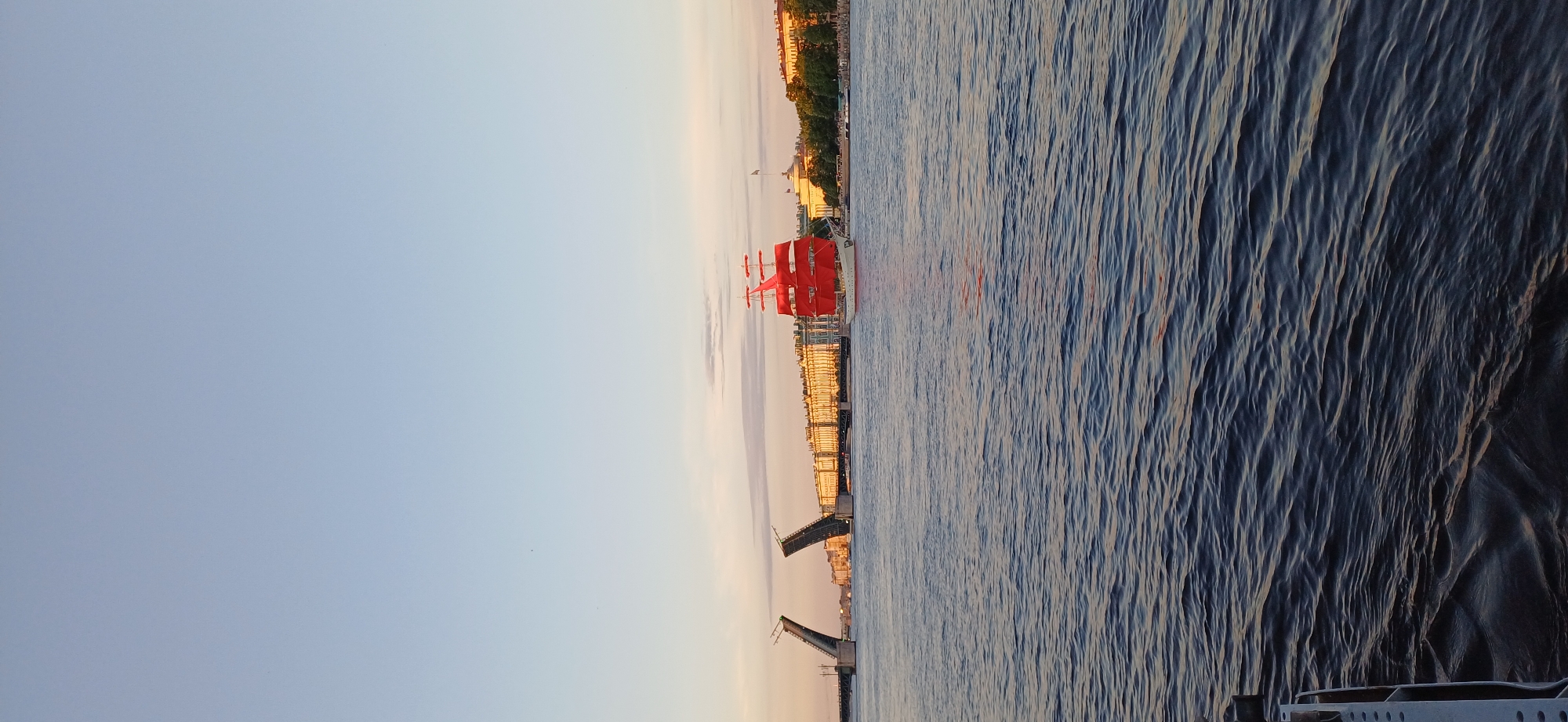 The image captures a view of the Neva River in Saint Petersburg, Russia, during sunset. In the foreground, dark blue water with small ripples stretches across the frame. In the mid-ground, a magnificent tall ship with bright red sails is sailing. The ship appears to be the "Krusenstern," a historic training vessel.
Behind the ship, the Palace Embankment (Dvortsovaya Naberezhnaya) is visible, lined with grand, cream-colored buildings. The Winter Palace is a prominent structure along this embankment. To the left, the drawbridges are raised, allowing passage for ships on the river. The sky above is a clear gradient of pale blue to light orange, indicating the sun is setting. The overall scene evokes a sense of grandeur and maritime tradition.